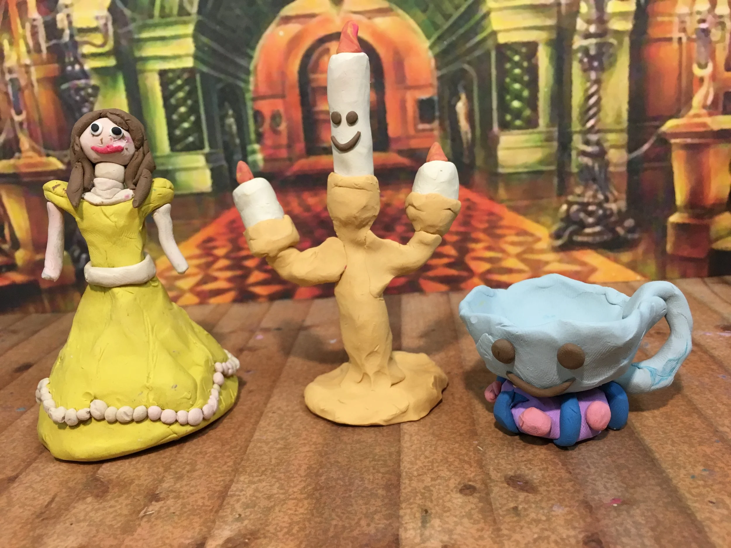 Clay figures of a woman in a yellow dress, a smiling candle, and a teacup with a face on a wooden surface with a colorful fantasy castle background.