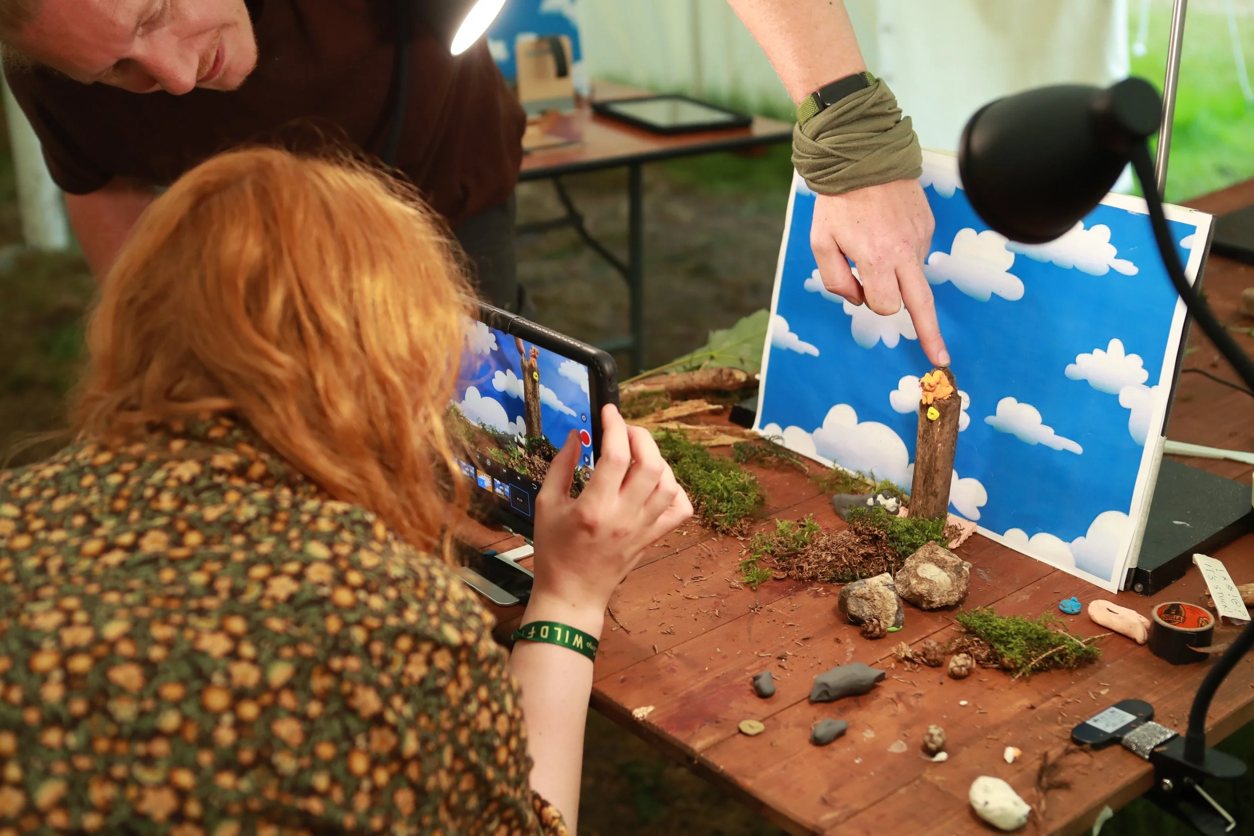 A person with a beard and dark shirt and a woman with red hair and a patterned shirt are showing a small diorama of a forest scene with rocks and moss, with a smiling face painted on a log, to a woman pointing at it. The woman is also recording the s