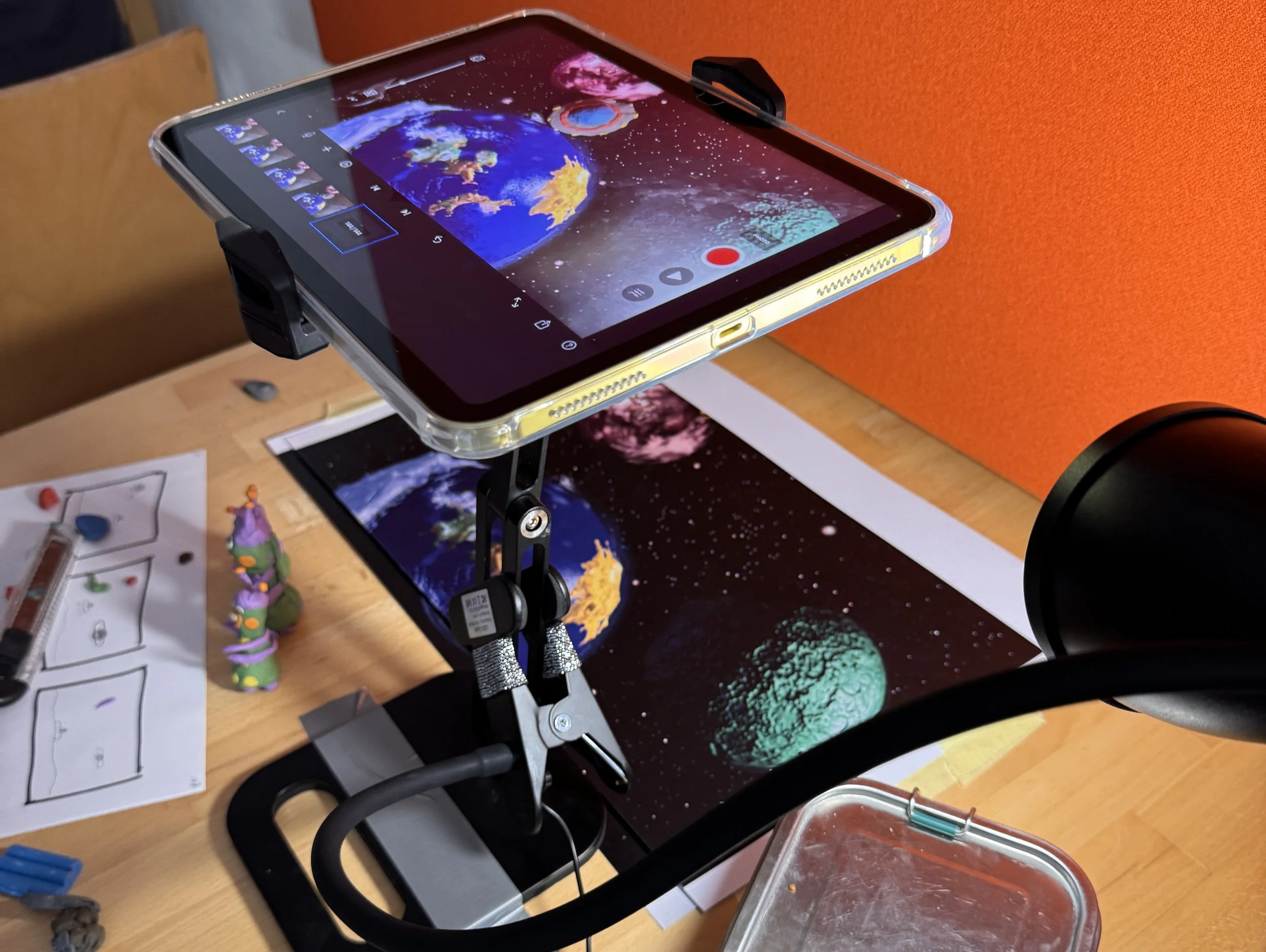 An orange lamp illuminating a workspace with a black telescope, a tablet, and a printed illustration. The tablet displays an image of Earth in space with planets and stars, mounted on a clamp on top of a desktop. The printed illustration shows Earth 