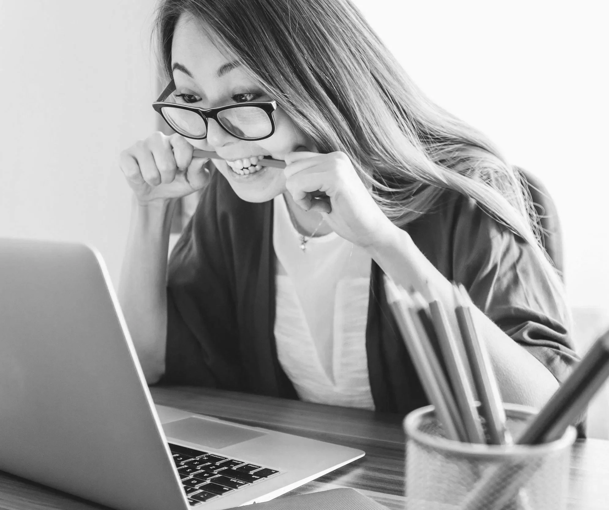 A woman with glasses biting her pencil while looking at her laptop in a workspace.