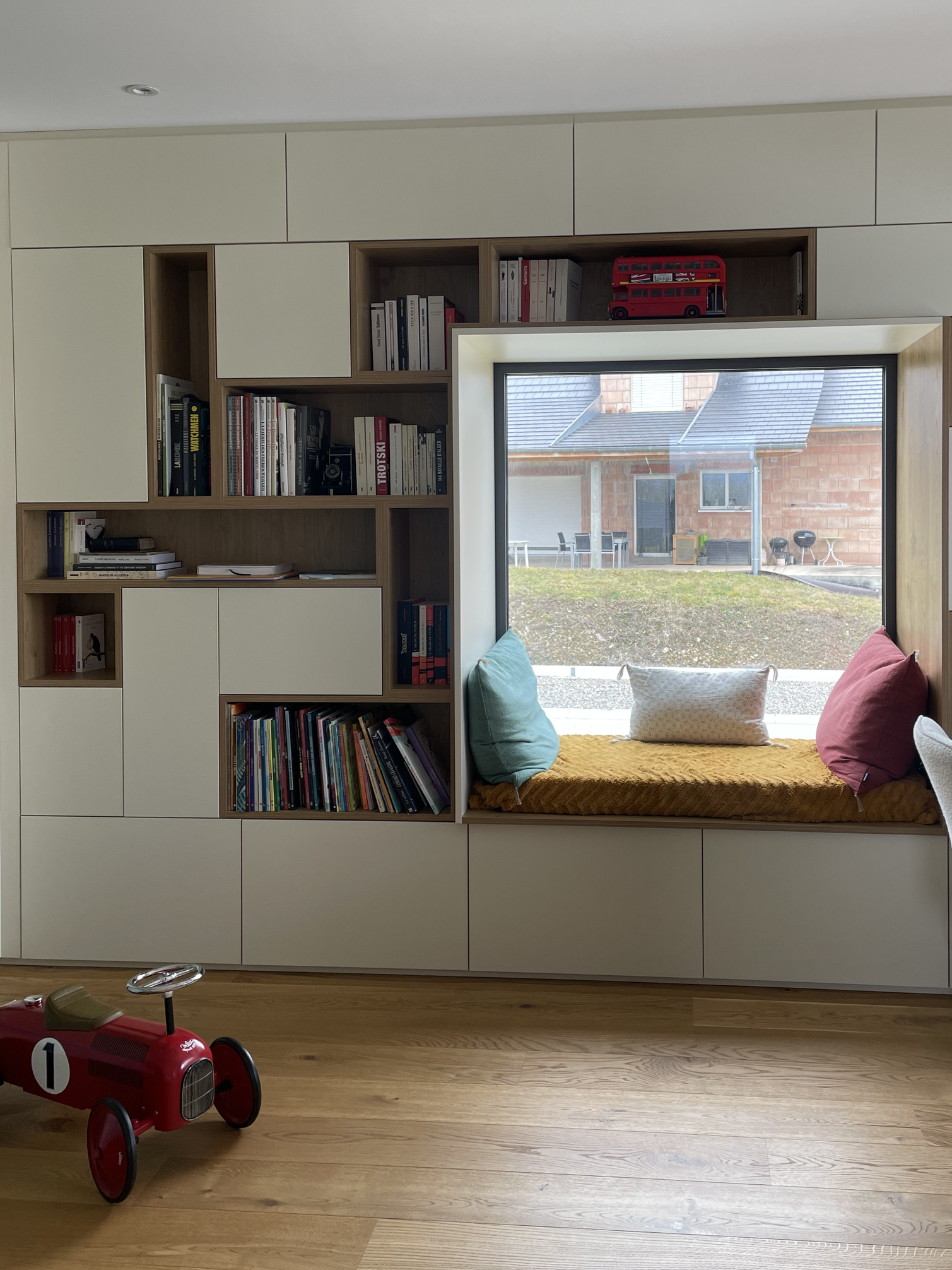 Bibliothèque asymétrique bois et crème avec banquette de lecture sous la fenêtre. Parquet aux sol et ambiance douce et lumineuse.
