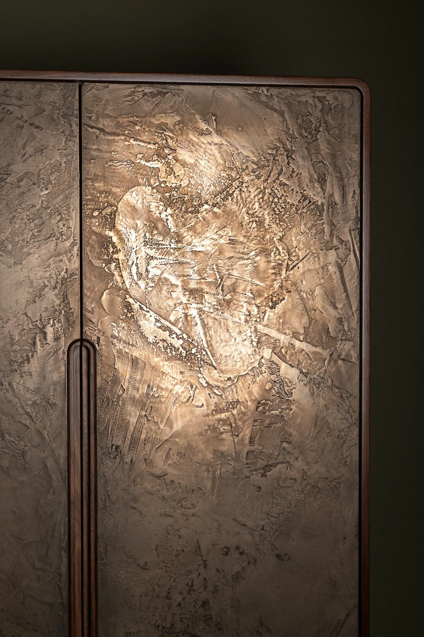Close-up of a bar cabinet with an aged bronze metallic surface with intricate, textured patterns and scratches. Cabinet has a solid Walnut case and solid walnut integral handle.