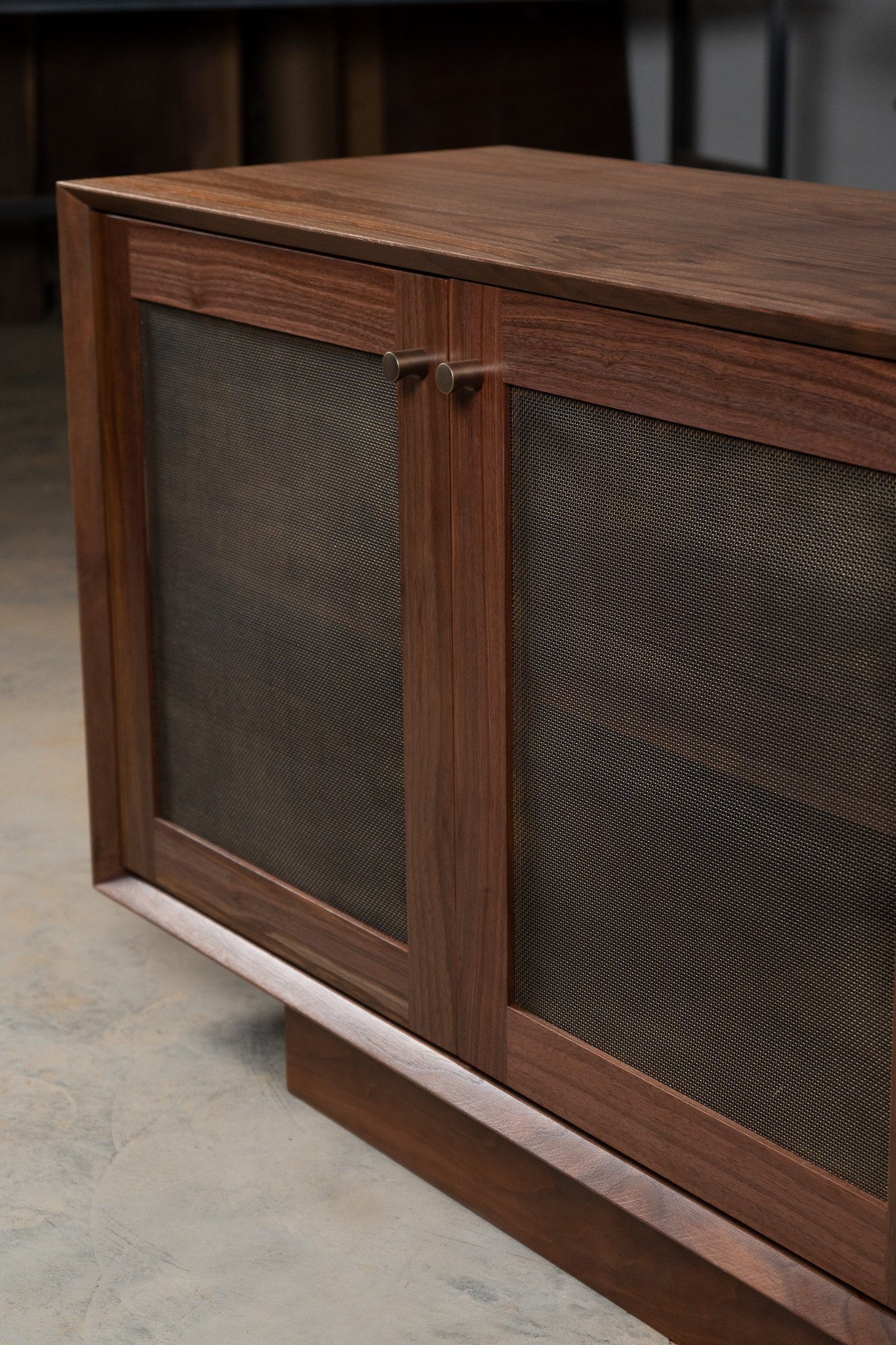 Close-up of a wooden cabinet with mesh speaker panels and round knobs, standing on a concrete floor in a room with dim lighting.