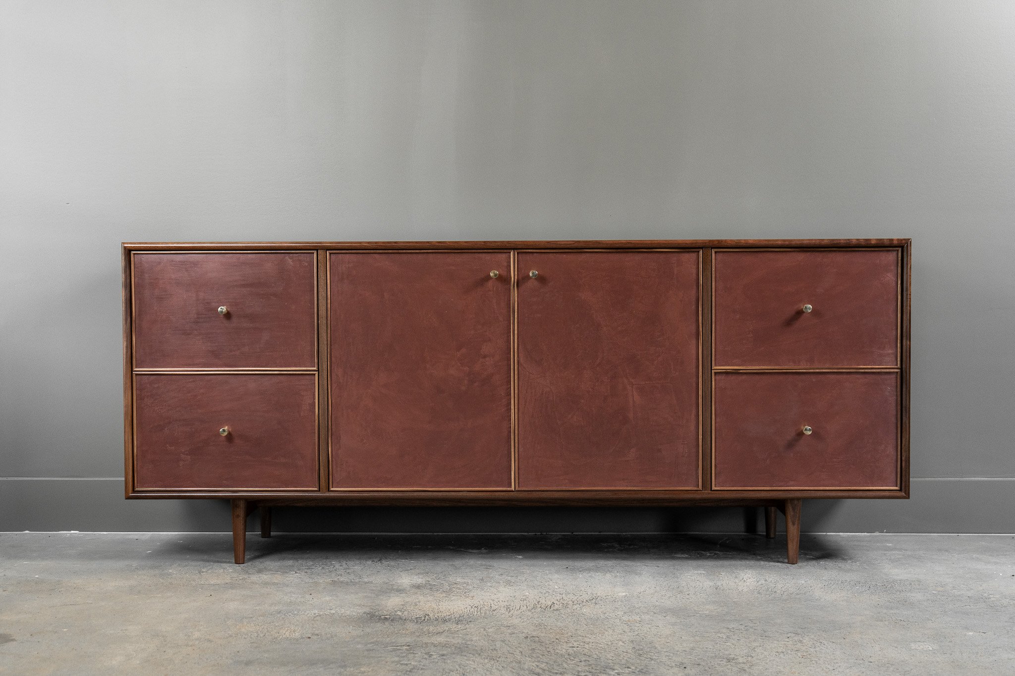 A handmade vintage modern sideboard cabinet with a medium burgundy hand troweled paster finish with beautiful texture visible.  four drawers and four doors with solid unlacuered brass knobs, standing against a gray wall on a concrete floor.