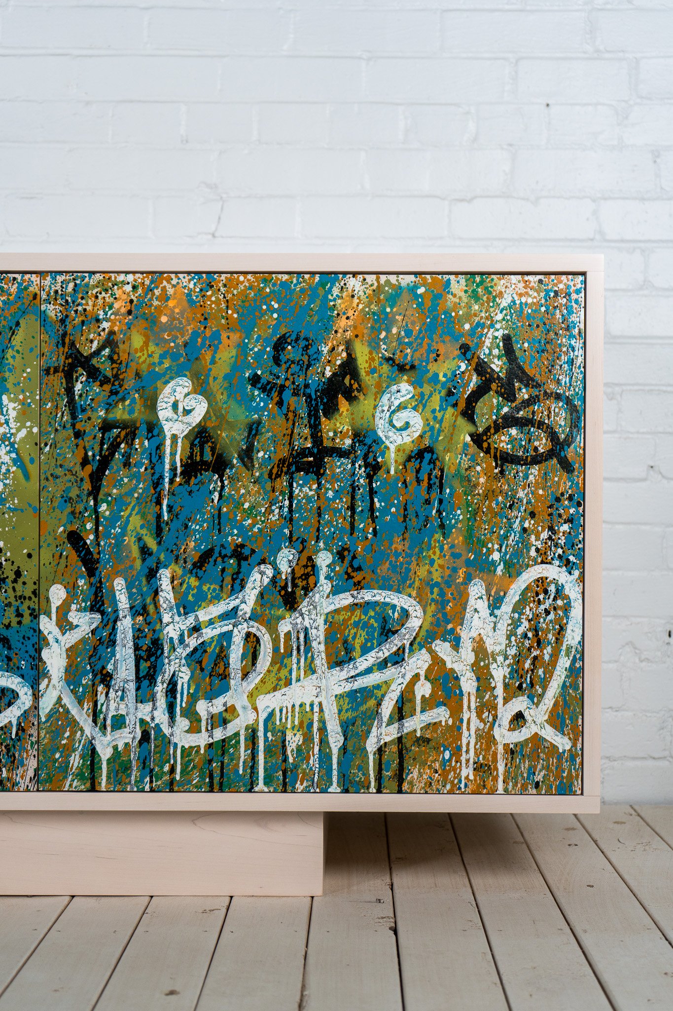 Close-up of a colorful abstract painted artwork with graffiti-style white writing on it, framed in a light wooden frame, placed against a white brick wall.