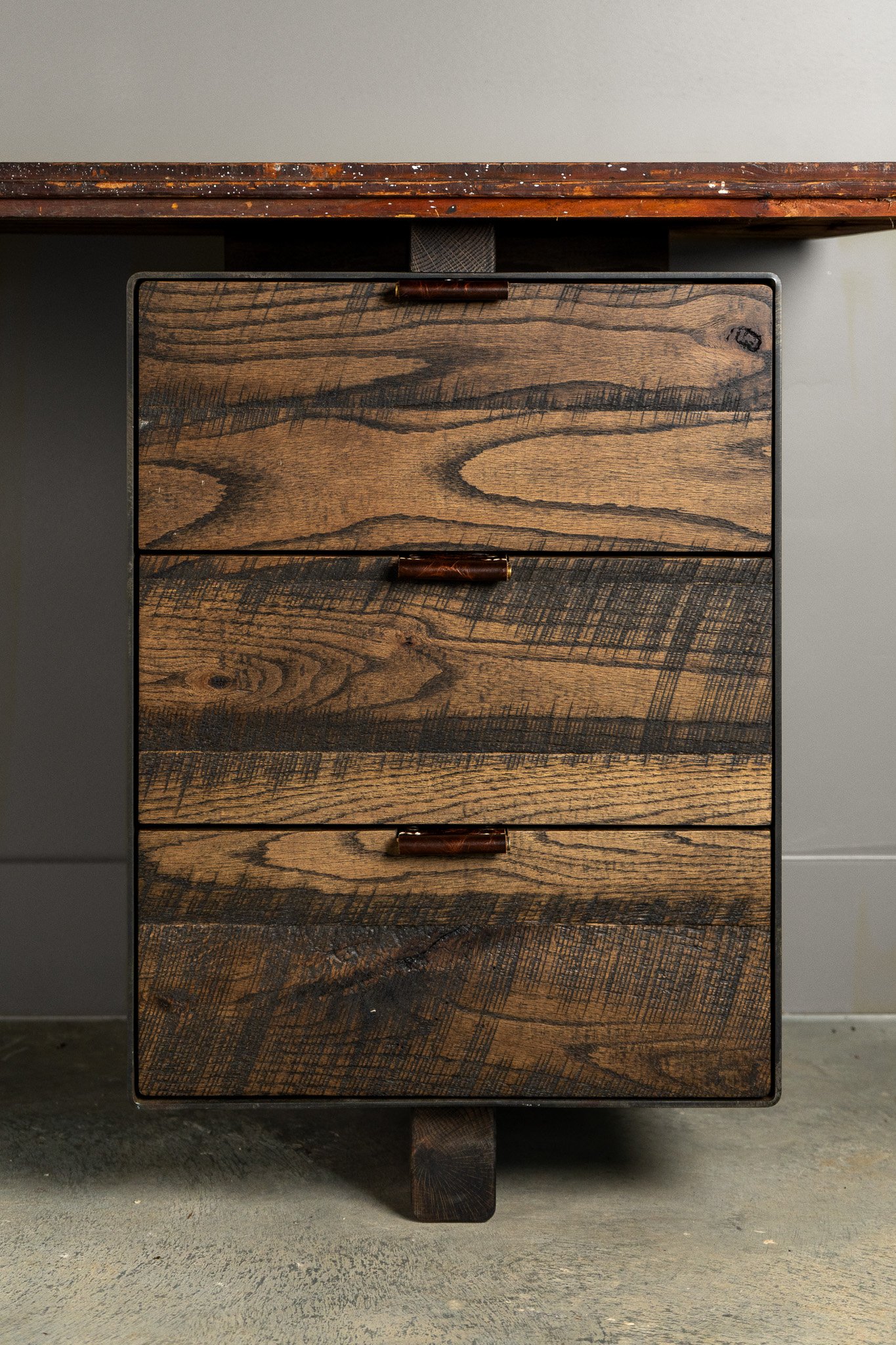 An organic modern desk drawer closeup with reclaimed wood for the front of the drawers with handmade leather pulls, positioned against a wall.