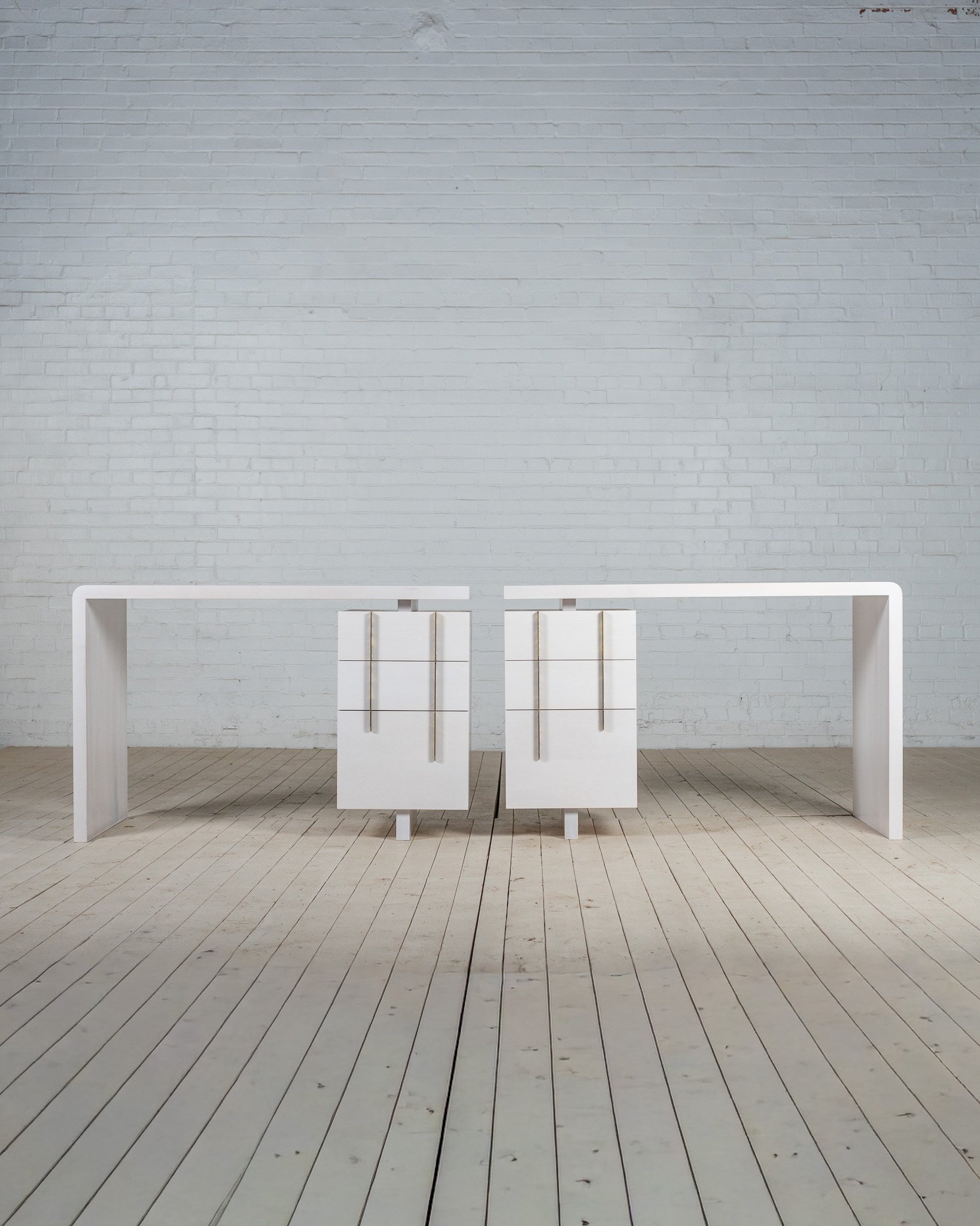 A large white room with white floorboards and a white brick wall background. In the center, three white sculptures or installations with vertical gold rods are placed on a white platform. A white table with curved legs is situated in front of the pla