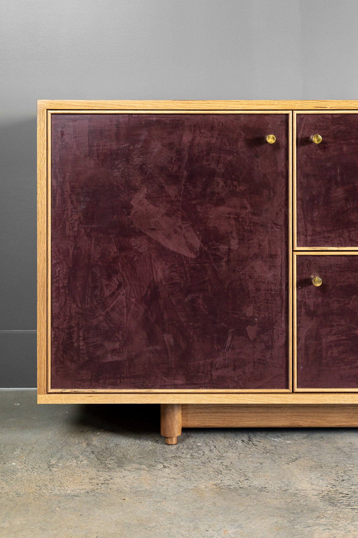 A mid-century modern solid white oak cabinet with a deep burgundy colored hand-troweled, textured finish along the front, and brass round knobs, standing on short rounded legs against a plain gray wall.