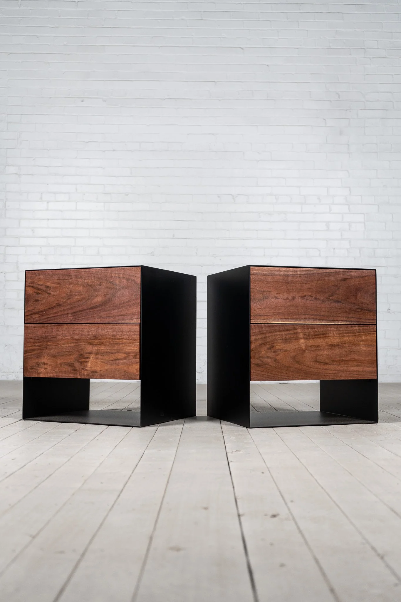 Two modern wooden cabinets with black sides are placed side by side in front of a white brick wall in a minimalistic room with white floorboards.