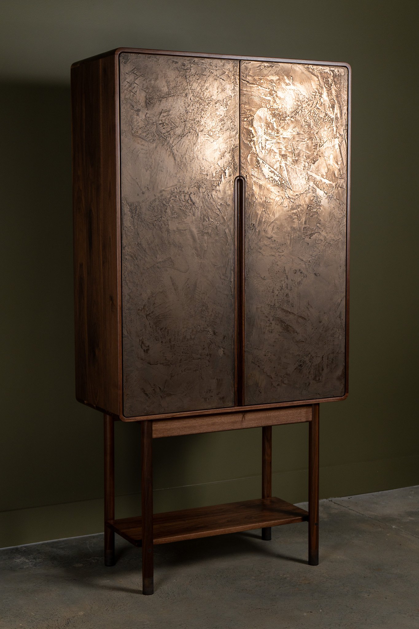 Wooden cabinet with textured metal doors on a stand against a green wall.