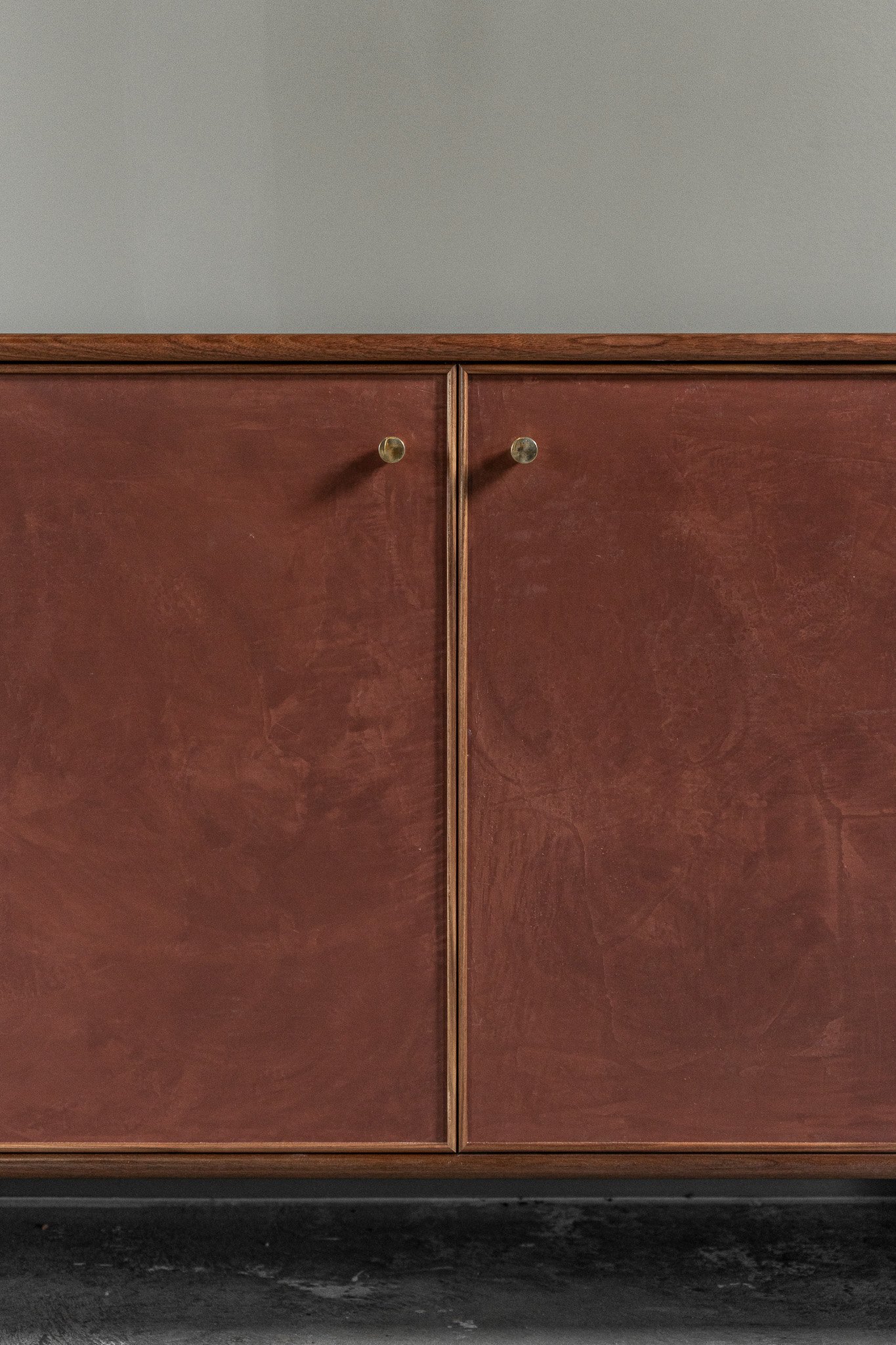 Close-up of a solid walnut cabinet with hand troweled plaster doors with a medium burgundy/red tone and beautiful texture visible. Doors have solid unlacquered brass knobs.