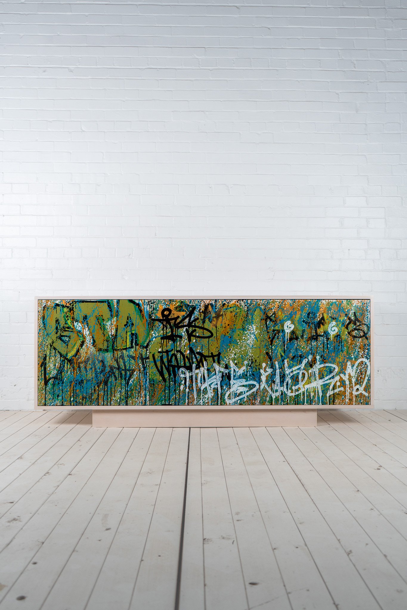 Solid maple handmade credenza with a white finish. Entire front of the cabinet is painted by a graffiti artist to look as though the cabinet has been vandalized with green, blue, white, orange, and black paint - on white floor with white brick wall.