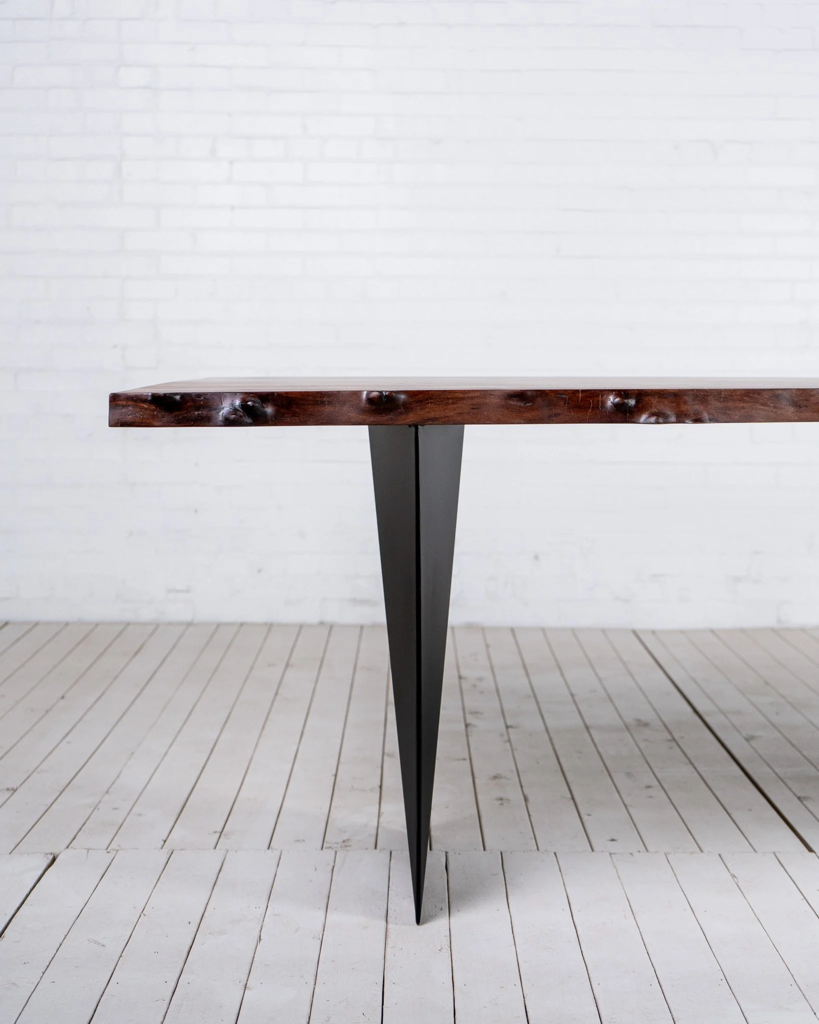 Close-up of a dark wooden tabletop with a black metal leg, set against a white brick wall and white wooden plank flooring.