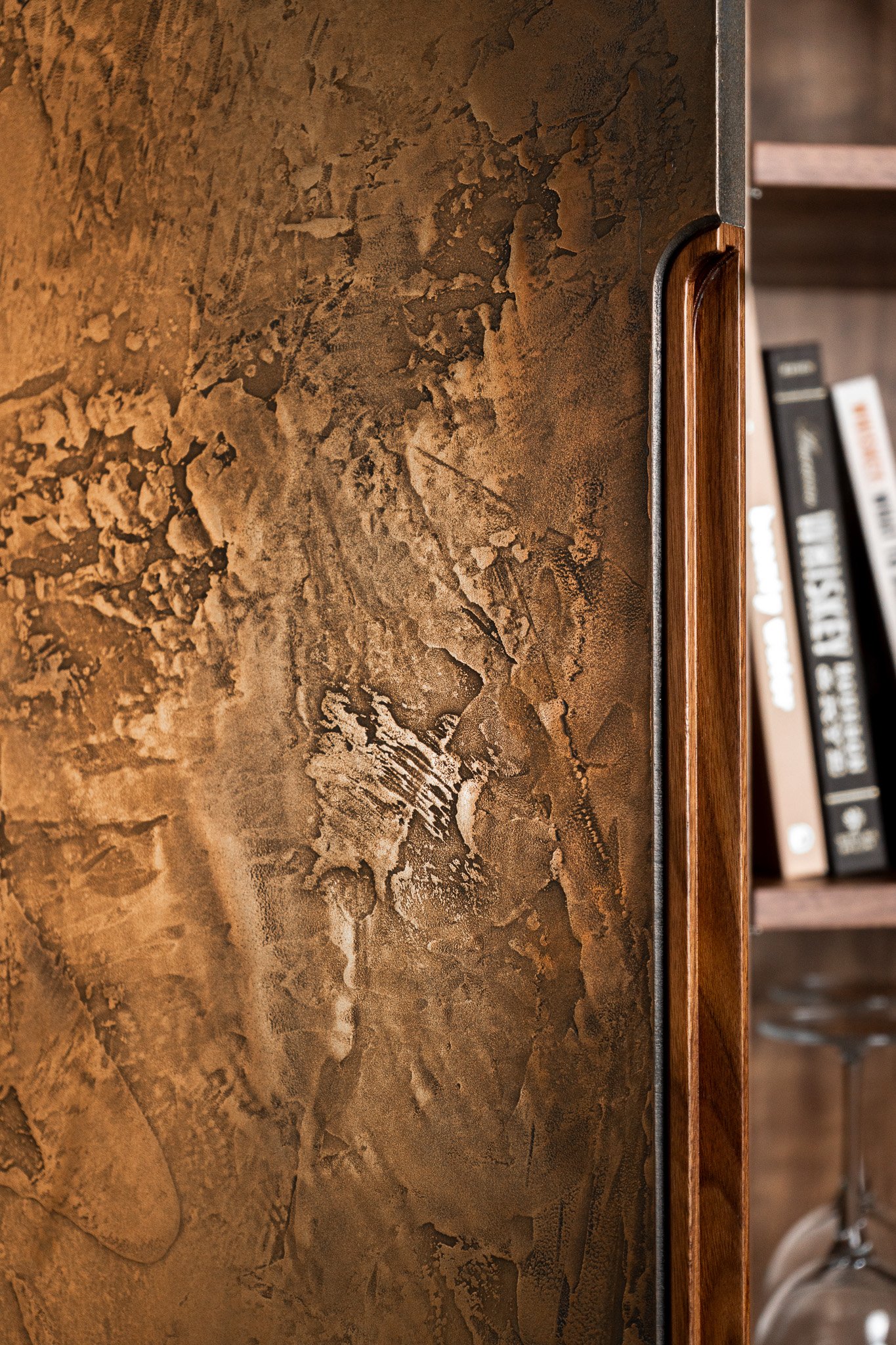 Close-up of a solid Walnut bar cabinet with a textured, metallic aged bronze door panel, showing an integral solid walnut handle on the right side. In the background, blurred books are visible on a shelf inside the cabinet.