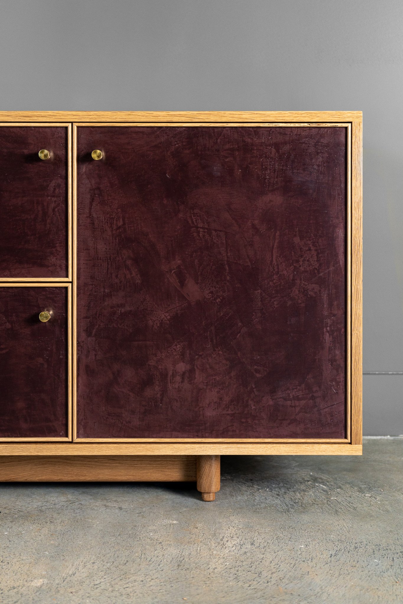 A mid-century modern wooden sideboard or cabinet with two large doors with a deep burgundy hand-troweled plaster textured finish, two smaller drawers with the same finish, against a plain gray wall, with a textured concrete floor.