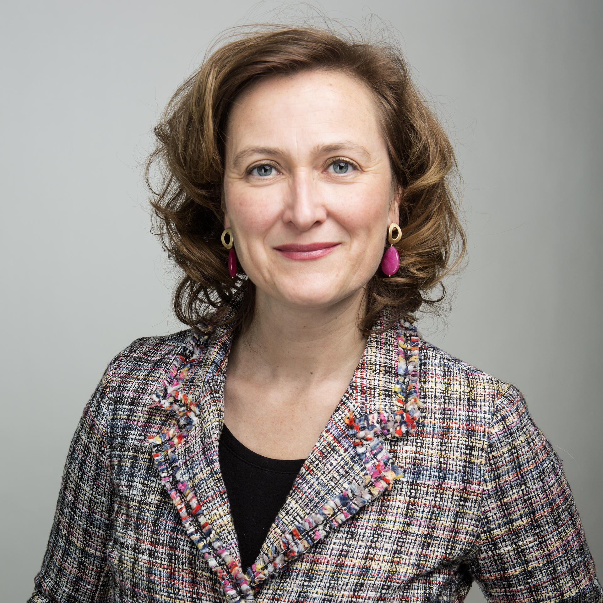 Een persoon met schouderlang, golvend haar draagt een kleurrijke, geruite blazer met een zwarte top eronder. Het gezicht van de persoon is geblurd en onherkenbaar gemaakt. De achtergrond is egaal grijs.