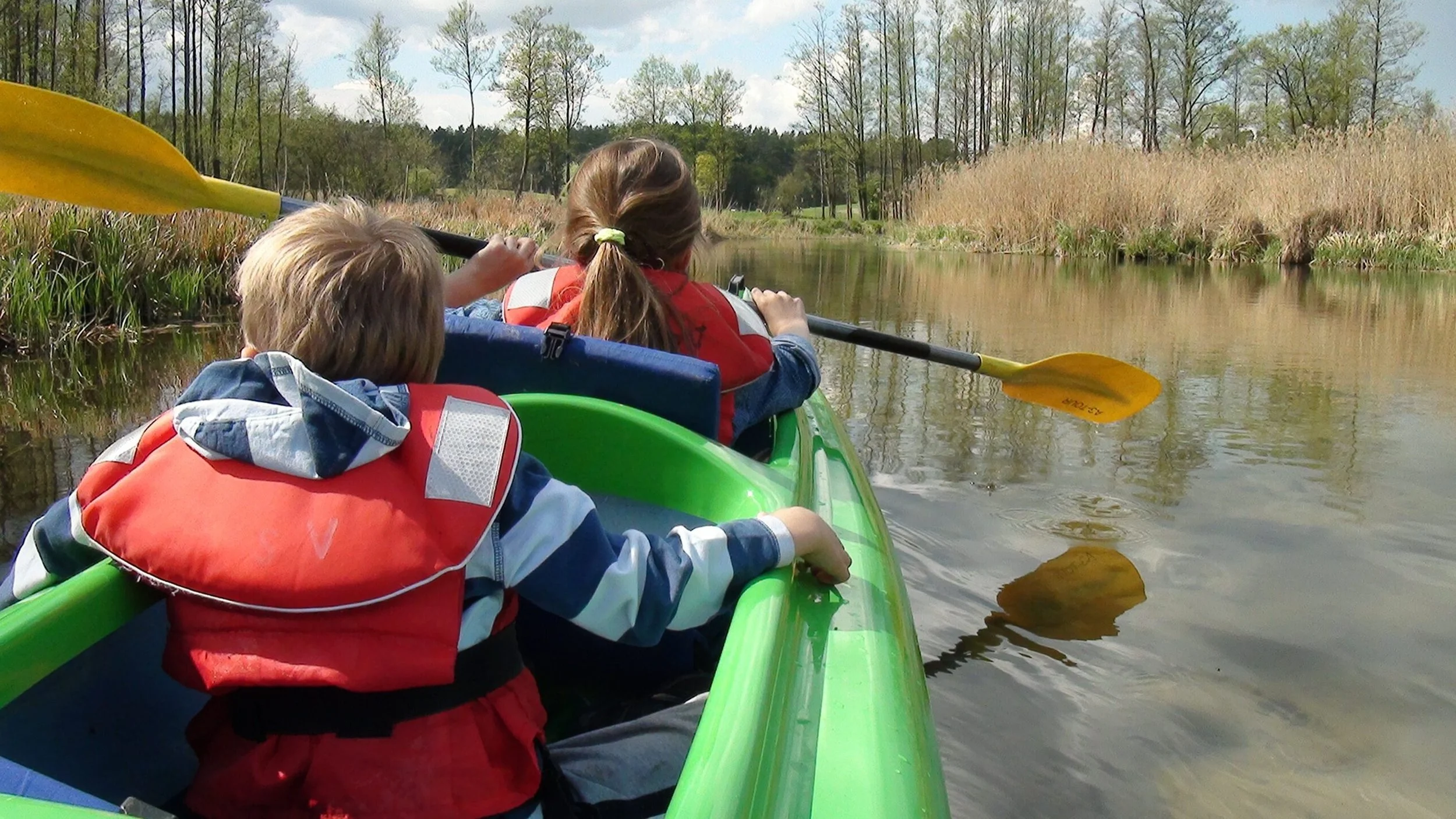 Kinderen in een groene kano varen op een rivier, omgeven door riet en bomen, onder een blauwe lucht.