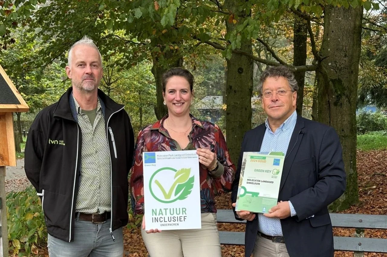 Drie personen poseren buiten met certificaten "Natuurinclusief" en "Green Key". Ze staan voor bomen, waarschijnlijk in een park of bos. Een van hen draagt een zwart jasje, een ander een kleurrijke blouse en de derde een donkerblauw colbert.