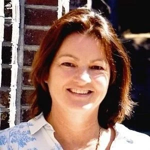 Vrouw met bruin haar glimlacht buiten voor een bakstenen muur.