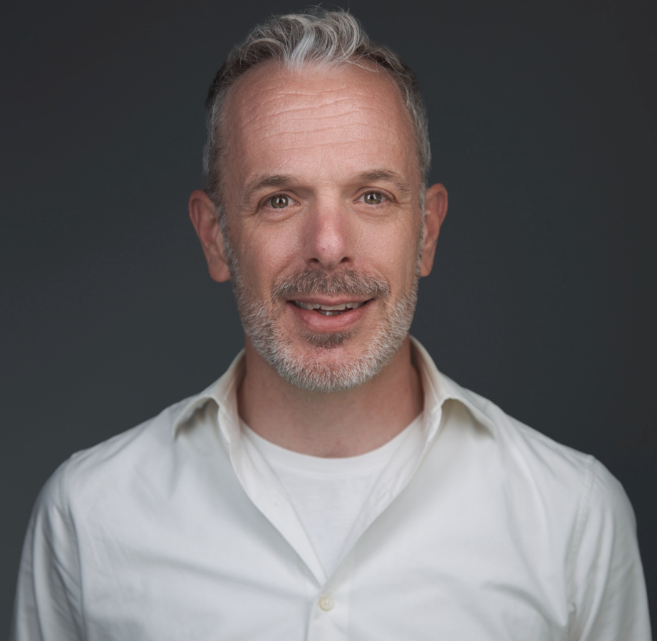 Portret van een lachende man met kort grijs haar en een baard, in een witte overhemd, tegen een donkere achtergrond.