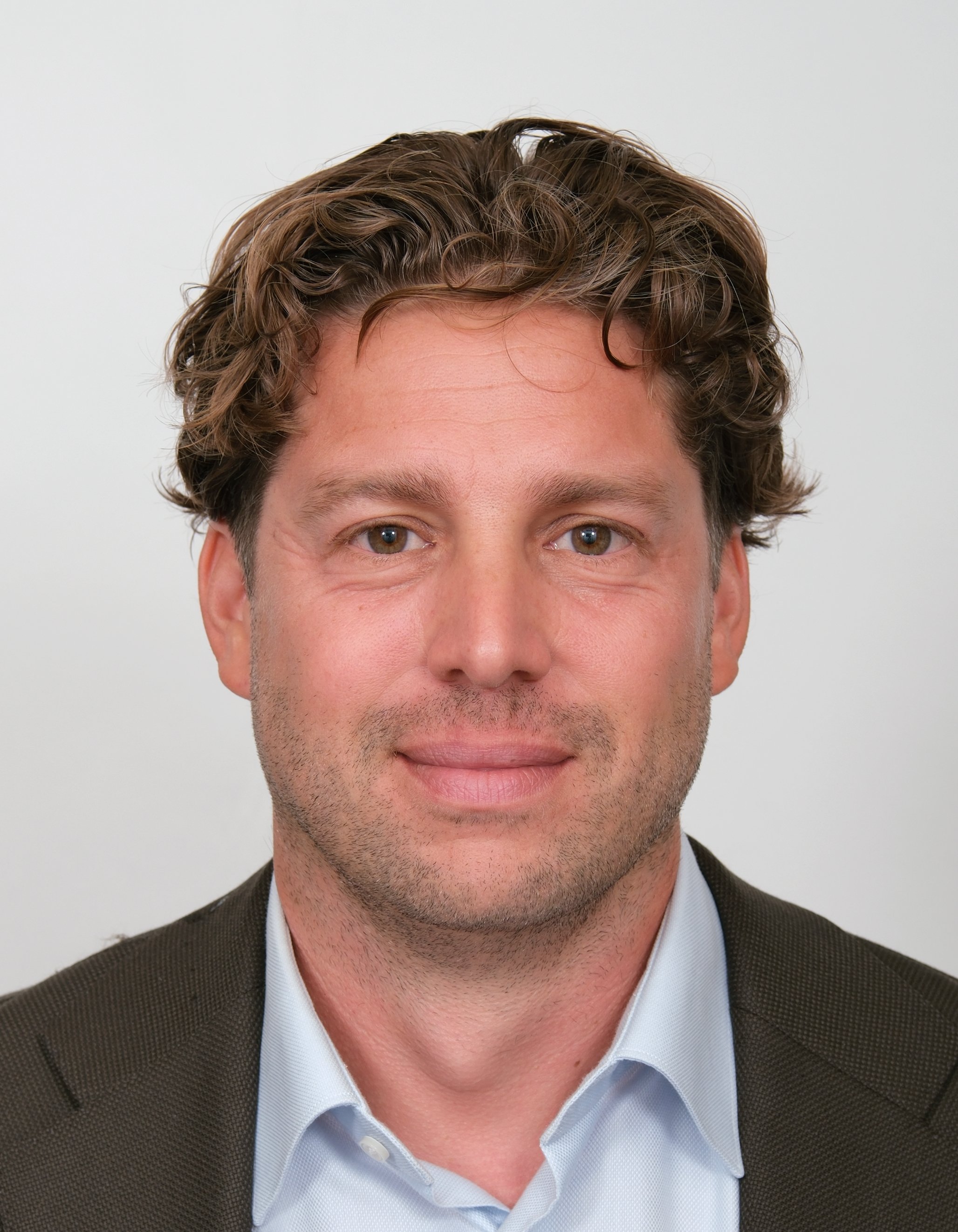 Man met donker haar glimlacht, draagt een colbert, blauwe achtergrond.