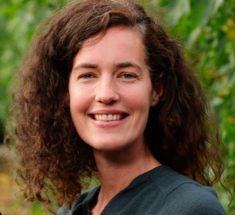 Portret van een vrouw met donker krullend haar, buitenomgeving met groene achtergrond.