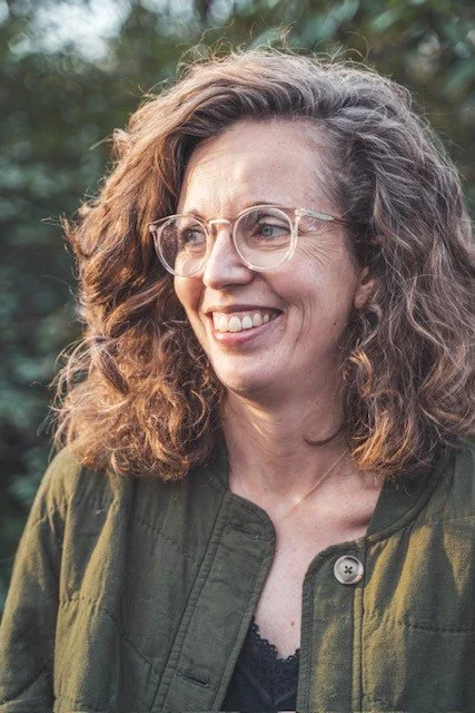 Vrouw met krullend haar en bril buiten in een groene omgeving.