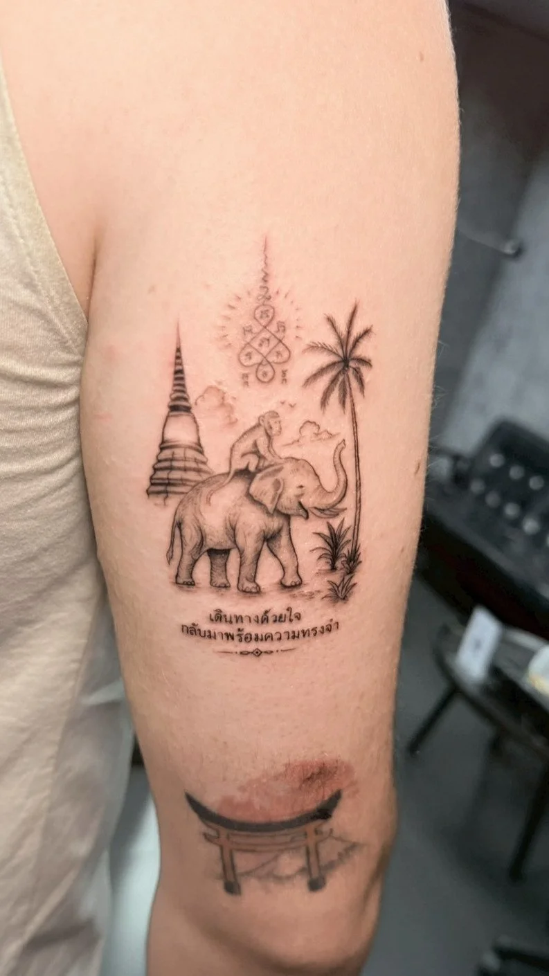 Tattoo of an elephant with a person sitting on its back, a palm tree, a stupa, a sacred symbol, and Thai script on a person's upper arm.