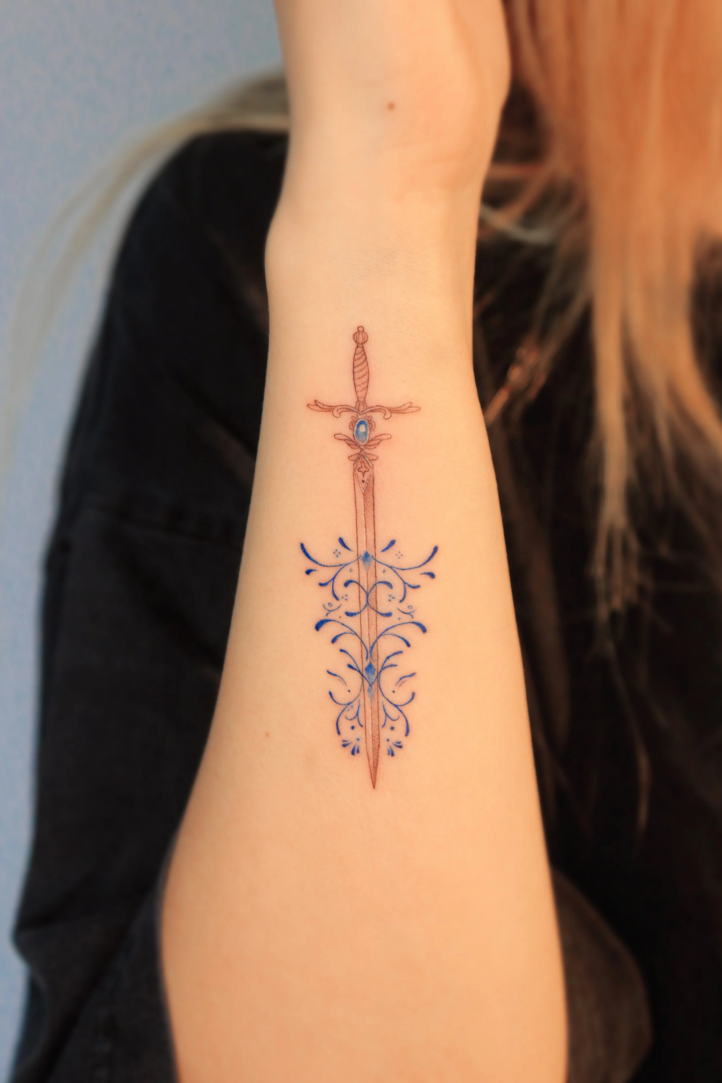 A tattoo of a sword with ornate blue ink decorations on the arm of a person in a black shirt.