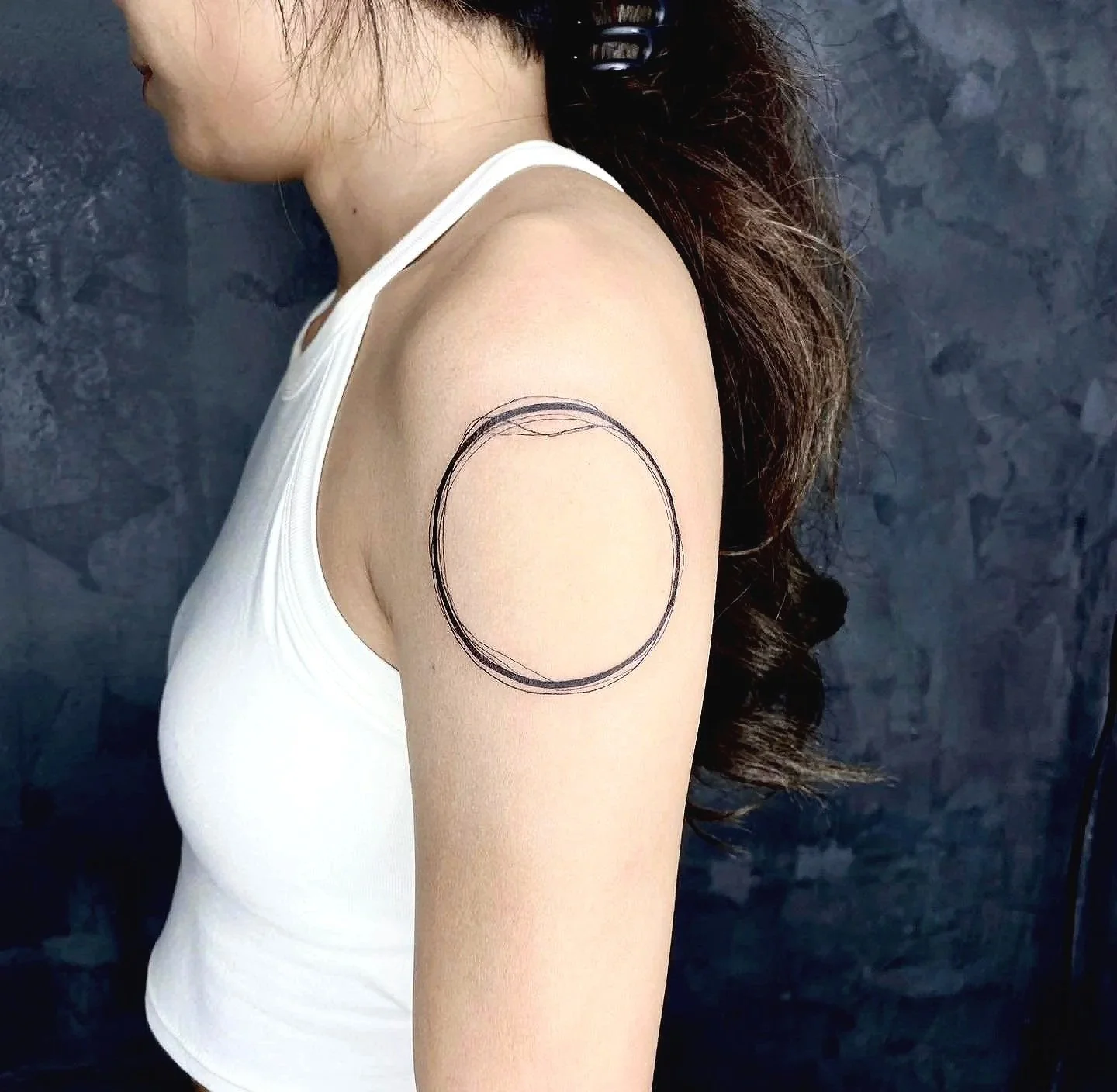A woman in a white tank top has a black ink circle sketch on her upper arm.