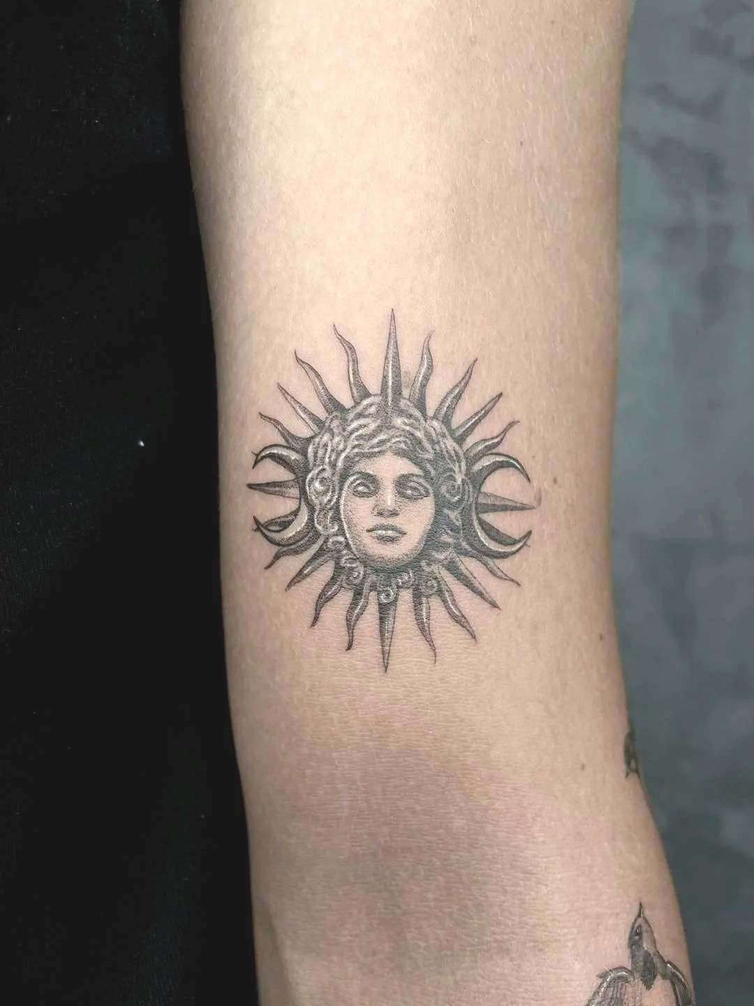 A tattoo of a woman's face with wavy hair, surrounded by radiating sun rays on a person's arm.