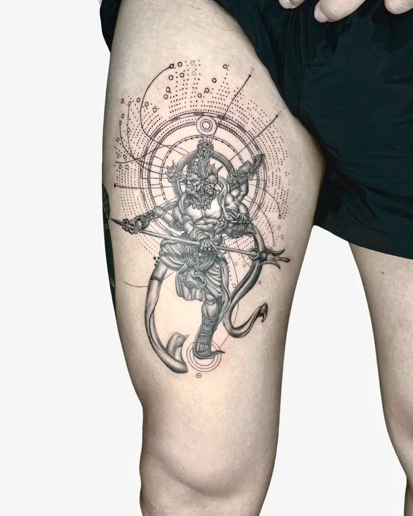 Black and gray tattoo of a fierce mythological or fantasy warrior with multiple limbs and a horned headdress, holding a spear and shield, with circular and geometric design elements surrounding the figure.