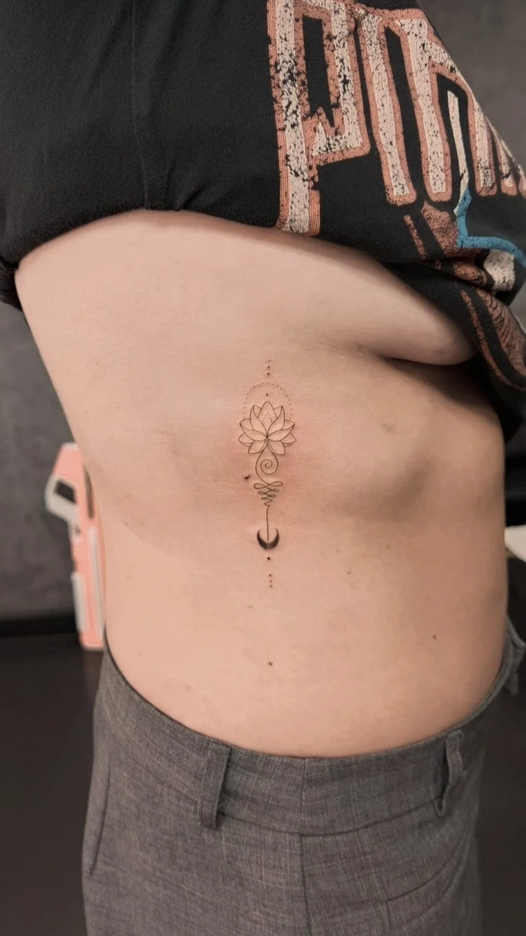 A tattoo on a person's side featuring a flower, a crescent moon, dots, and the word 'Love' in cursive