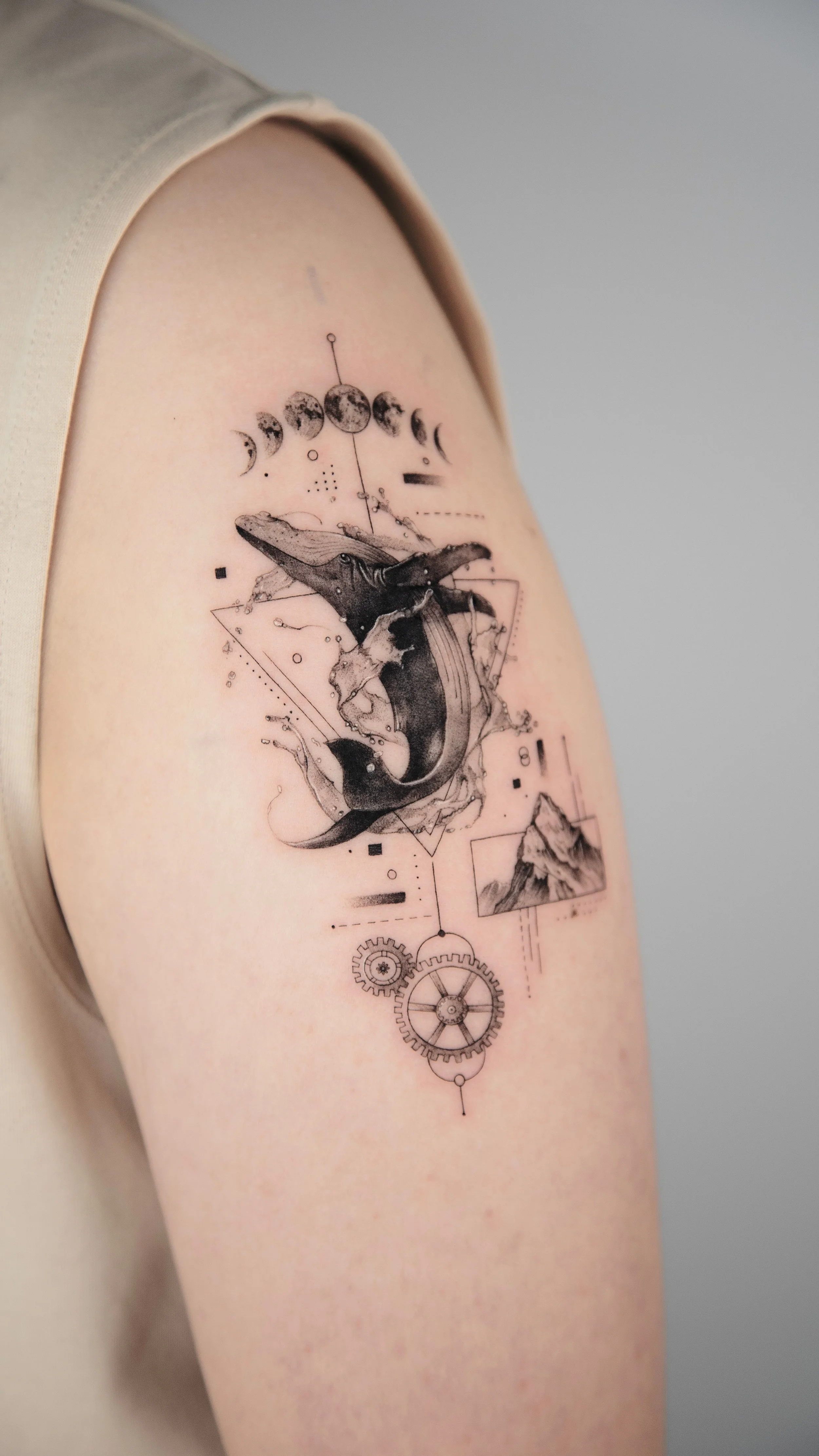 Tattoo of a whale jumping out of the water with moon phases and geometric and mechanical elements around it on person's upper arm.