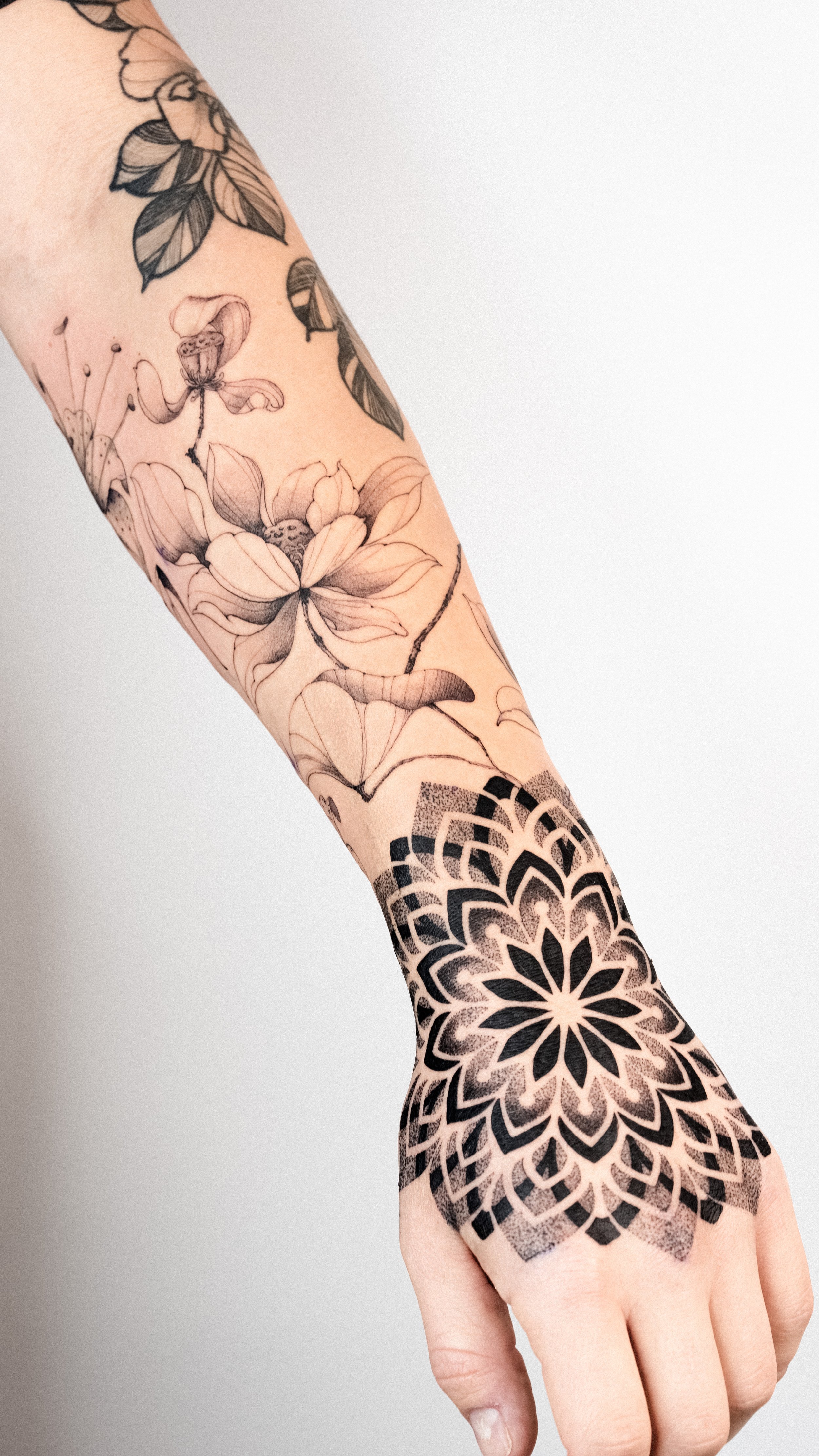 Close-up of a person's arm and hand with floral and geometric black ink tattoos, including detailed flowers and a mandala pattern.