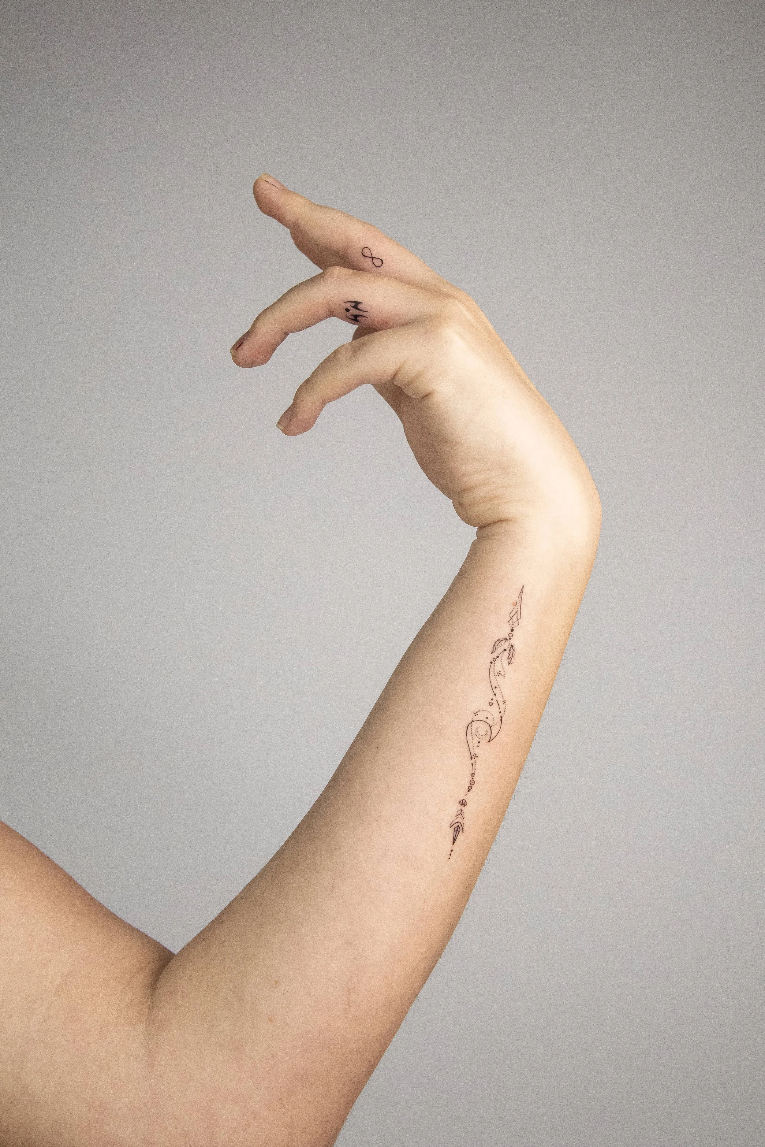 A human arm with a tattoo of a delicate, abstract design running along the inner forearm. The arm is held up and slightly bent, with the hand's fingers partially curled, against a plain light gray background.
