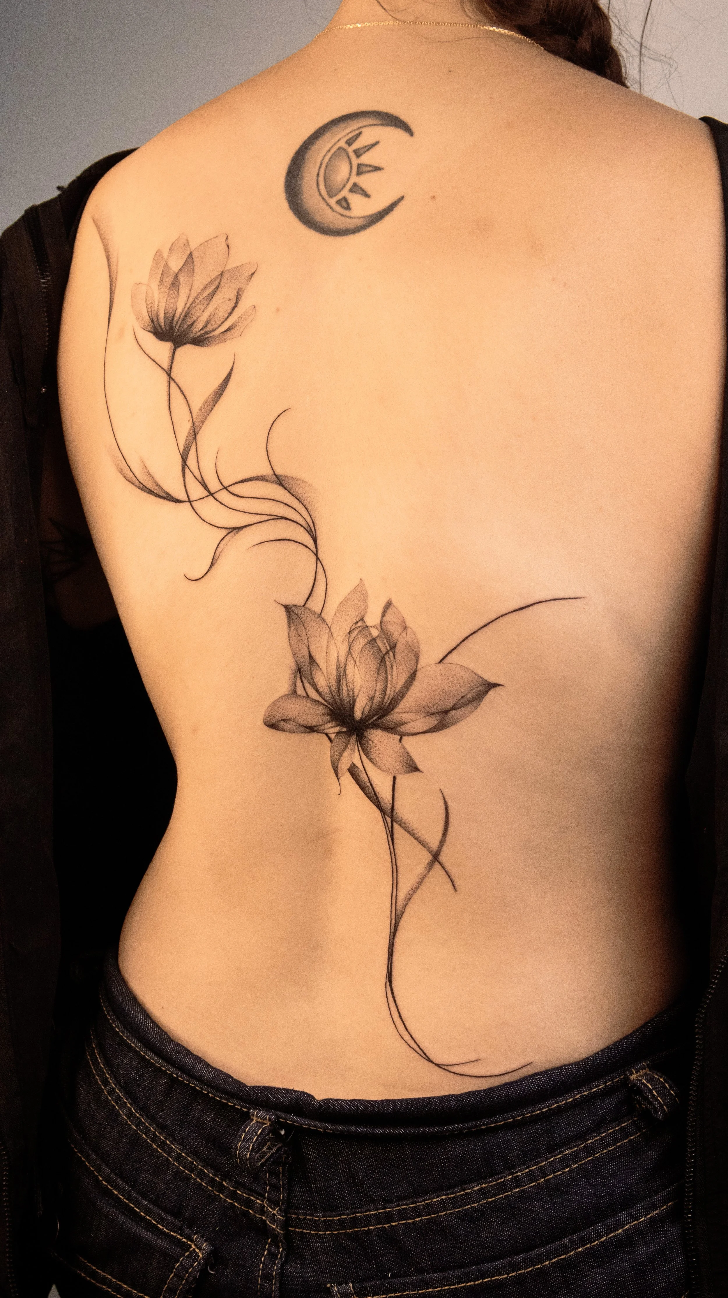 Black ink tattoo of two blooming flowers connected by flowing lines on a person's upper back and shoulder area, with a crescent moon and sun symbol tattoo above the flowers.