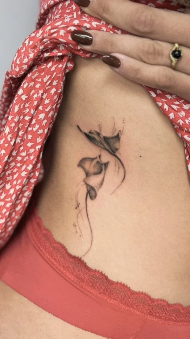 A tattoo of a butterfly on a woman's side, near the waist, with part of her hand visible pulling up her shirt, showing a ring on her ring finger, and wearing a red garment with white leaf patterns.