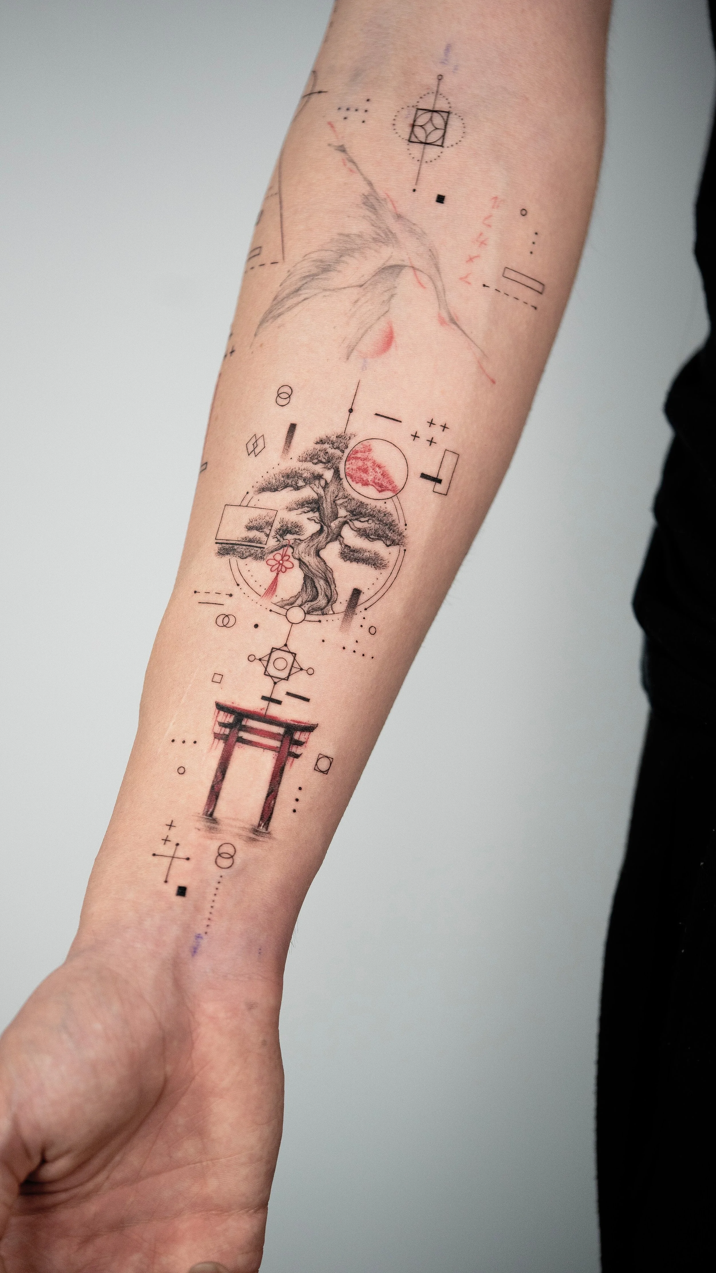 Tattoo on forearm featuring a bonsai tree, a red circle, geometric shapes, and a traditional Japanese torii gate, combined with abstract lines and dots.