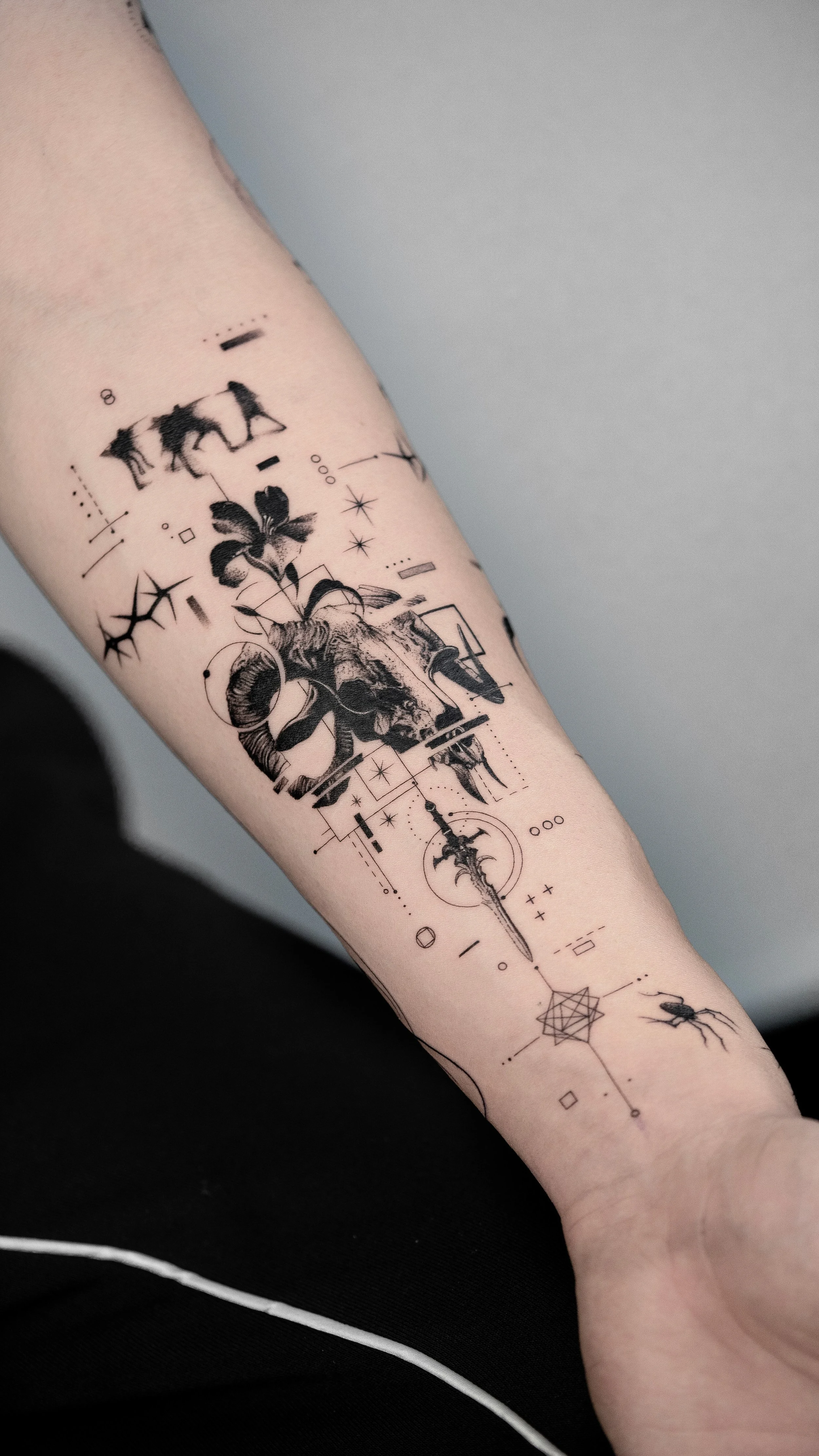 A person's forearm tattoo with various geometric shapes, a butterfly, a skull, a sword, insects, and decorative elements in black ink.