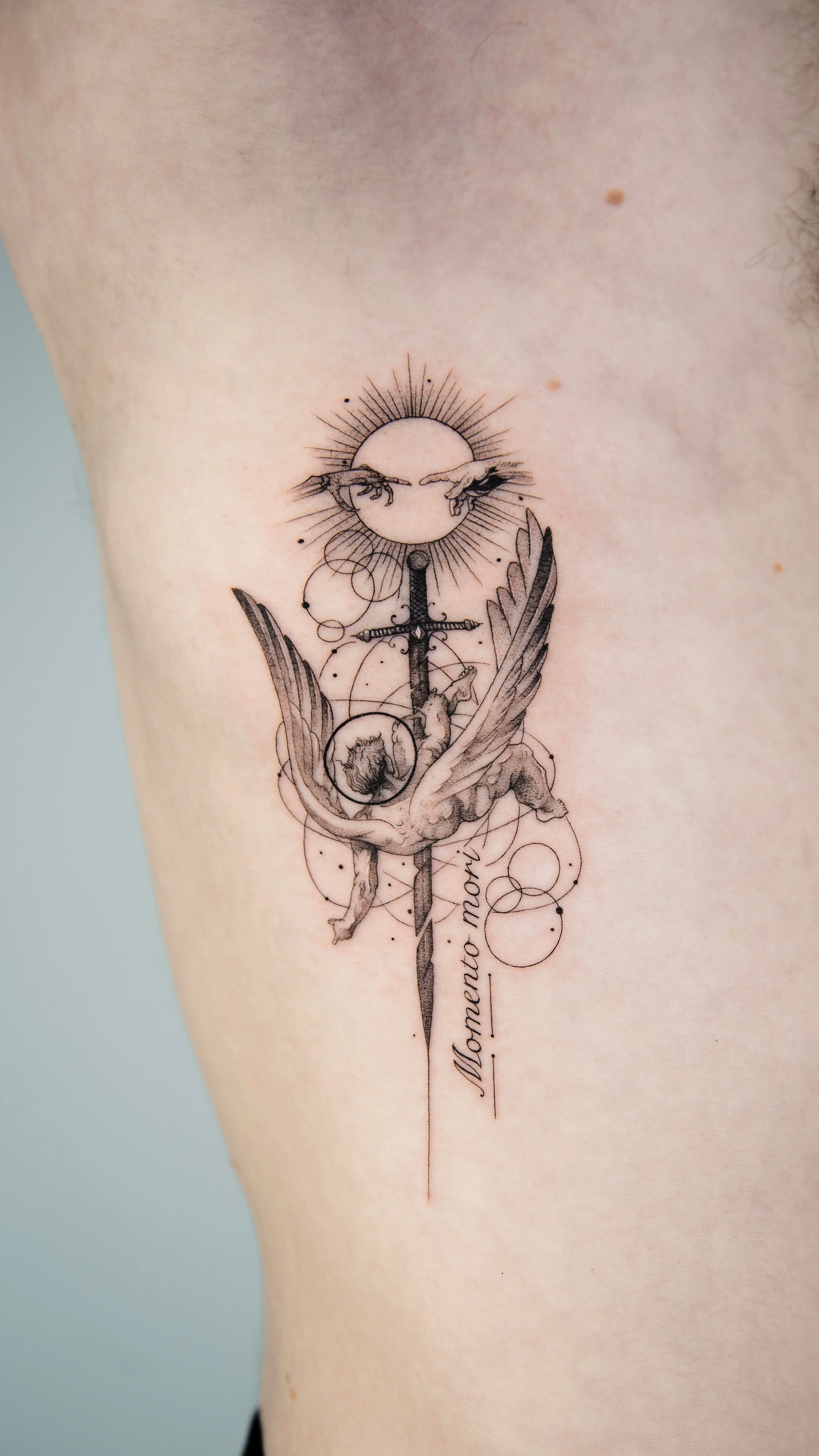 Tattoo design featuring a sun, two hands reaching toward each other, a winged figure, a sword, and Spanish text 'Memento Mori' on the person's upper arm.