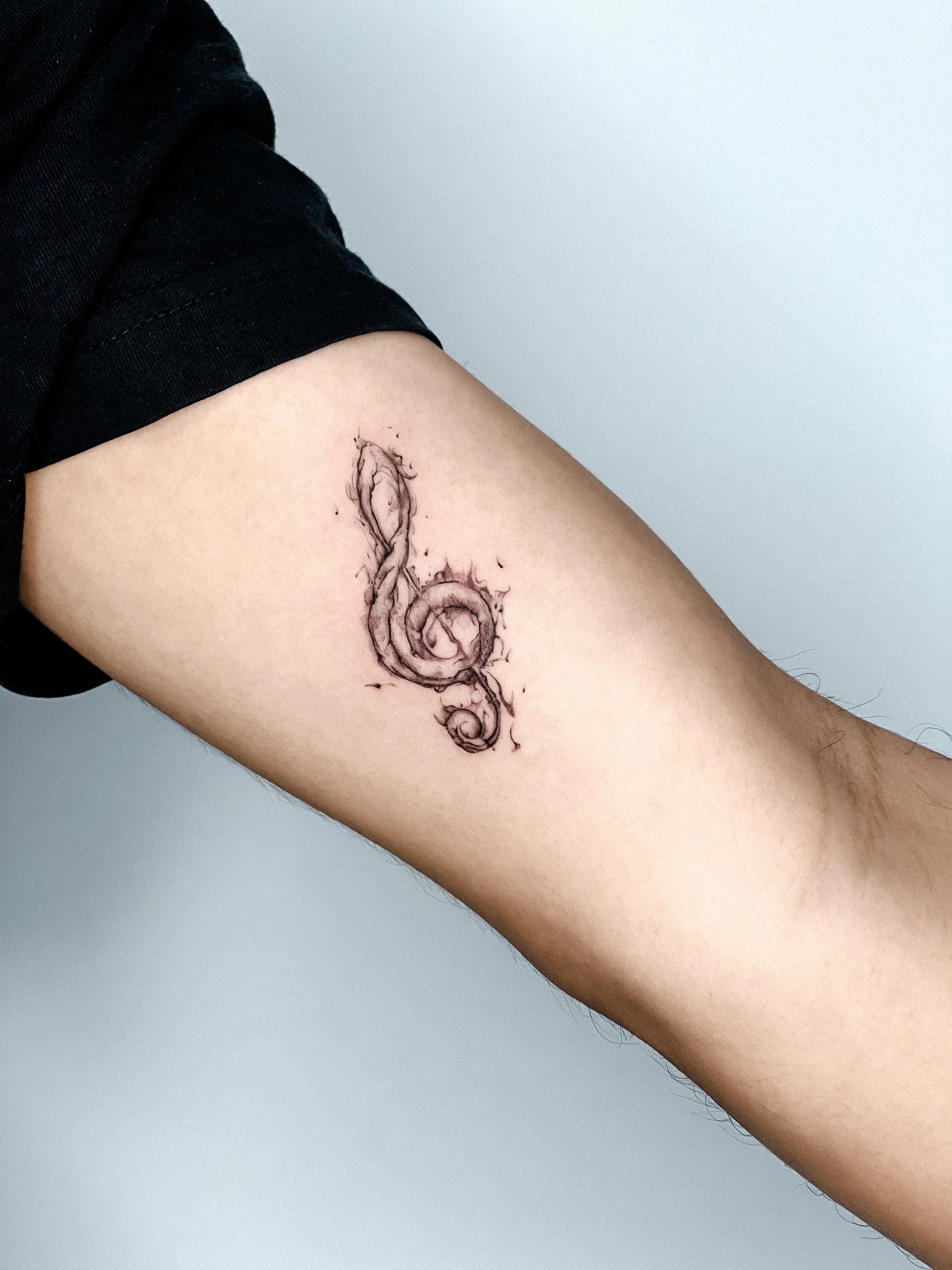 Tattoo of an abstract, flowing design in black ink on a person's upper arm, partially covered by a black shirt sleeve.