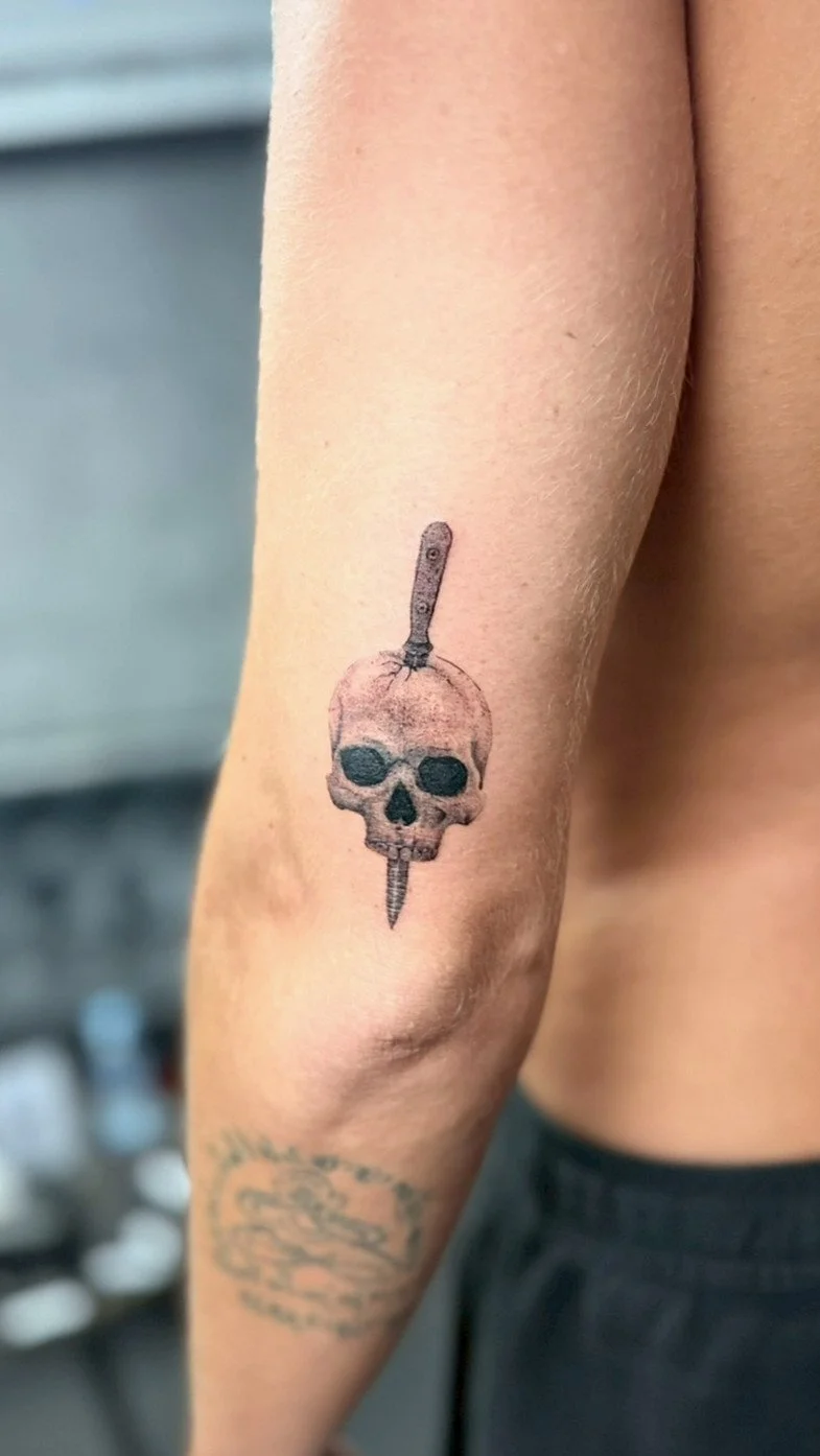 Tattoo of a skull with a knife through the top and bottom of the skull on a person's arm.