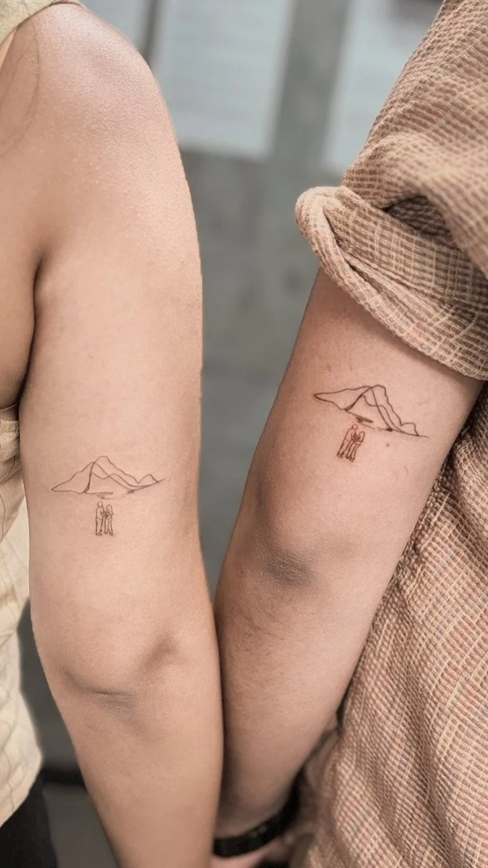 Close-up of two people's upper arms with matching minimalist tattoos of mountain ranges and two figures standing beneath a cloud