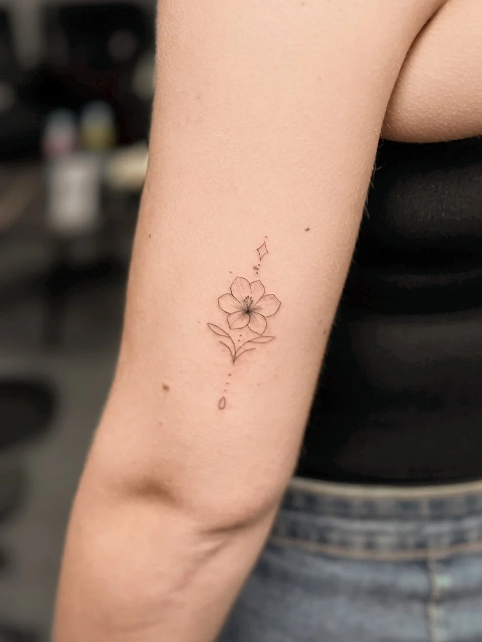 Delicate black ink tattoo of a flower on a person's inner forearm with small decorative elements and the word 'love' written vertically.