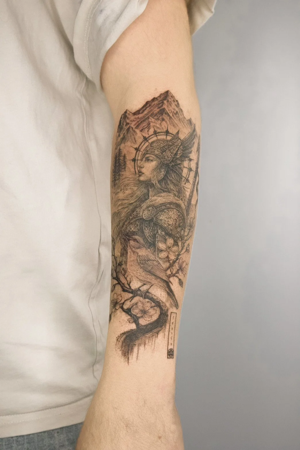 A tattoo on a person's arm featuring a Native American woman in traditional headdress, a mountain landscape, a bird perched on a flowering branch, and a small rectangular symbol at the bottom.