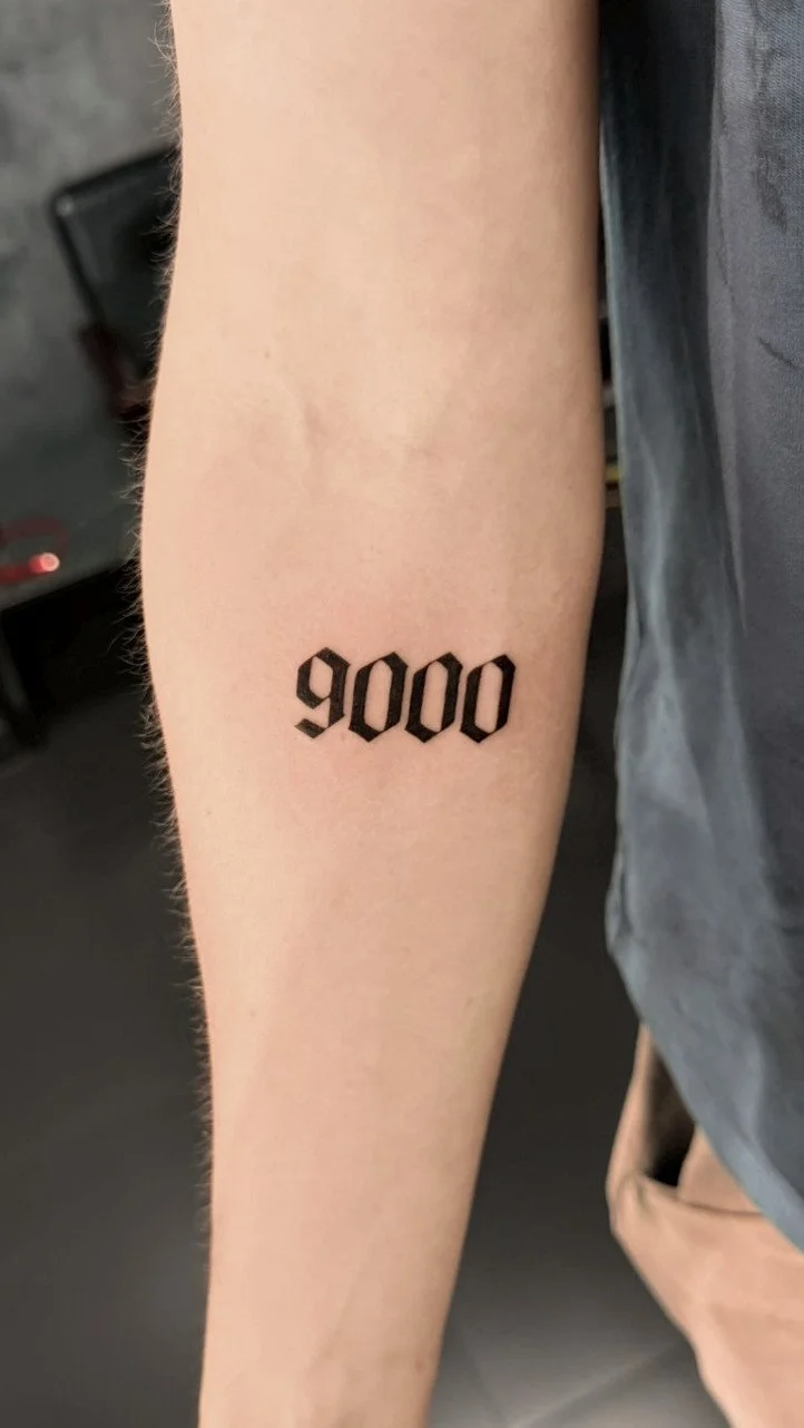 Tattoo of the number 9000 in black gothic font on a person's forearm.