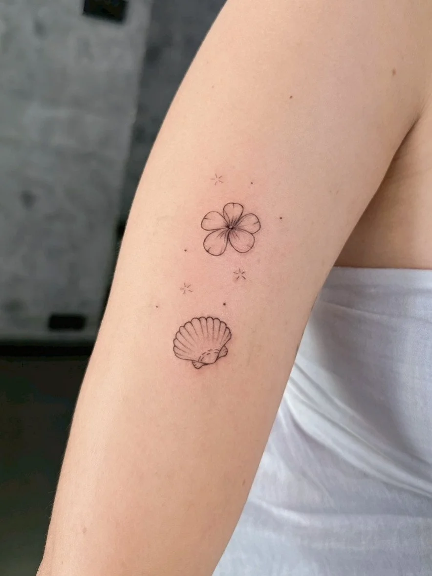 A tattoo of a flower and a seashell on a person's upper arm, with small decorative stars surrounding them.