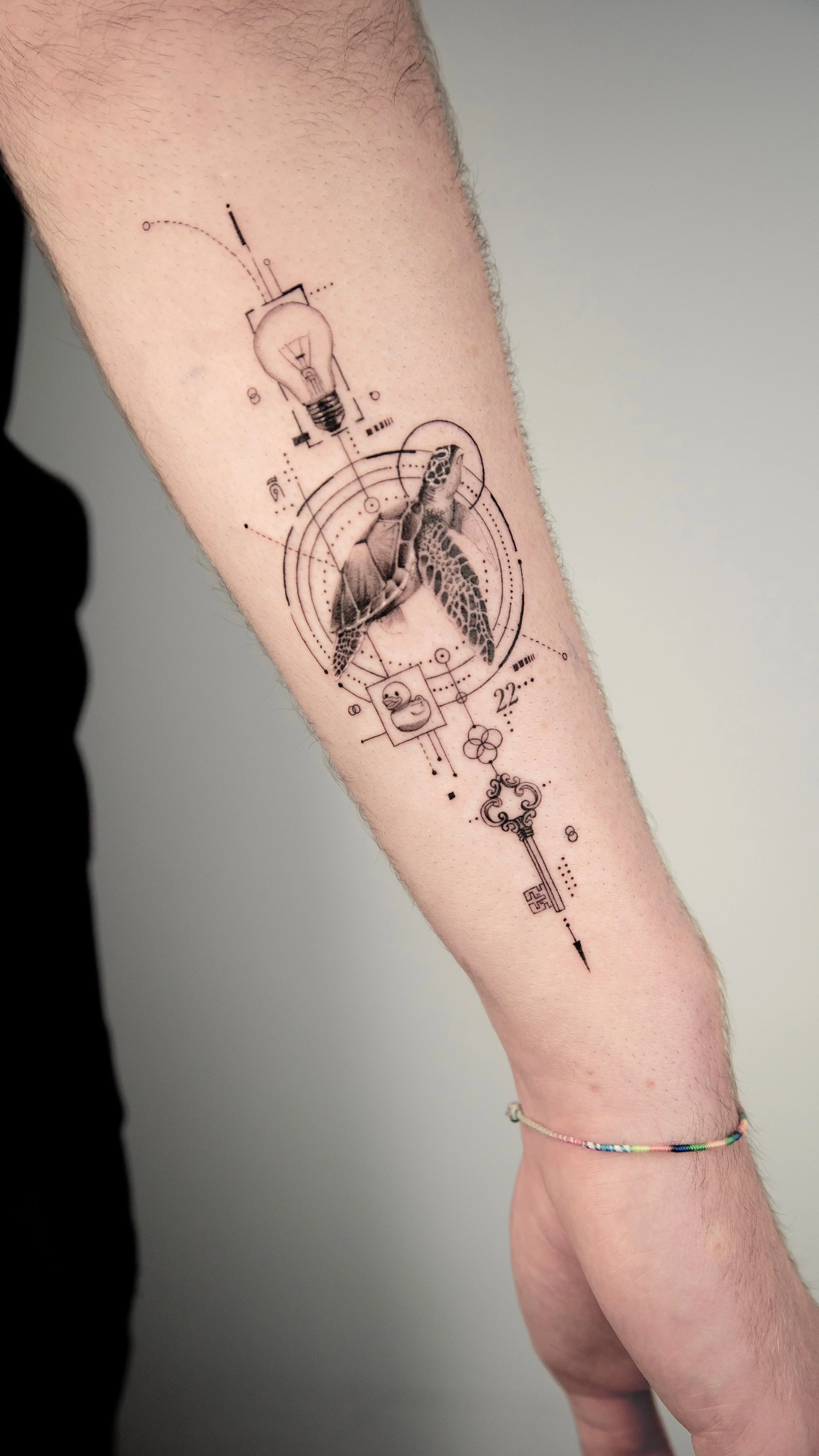 Tattoo of a turtle, an abstract key, and a light bulb with geometric and technical design elements on a person's forearm.