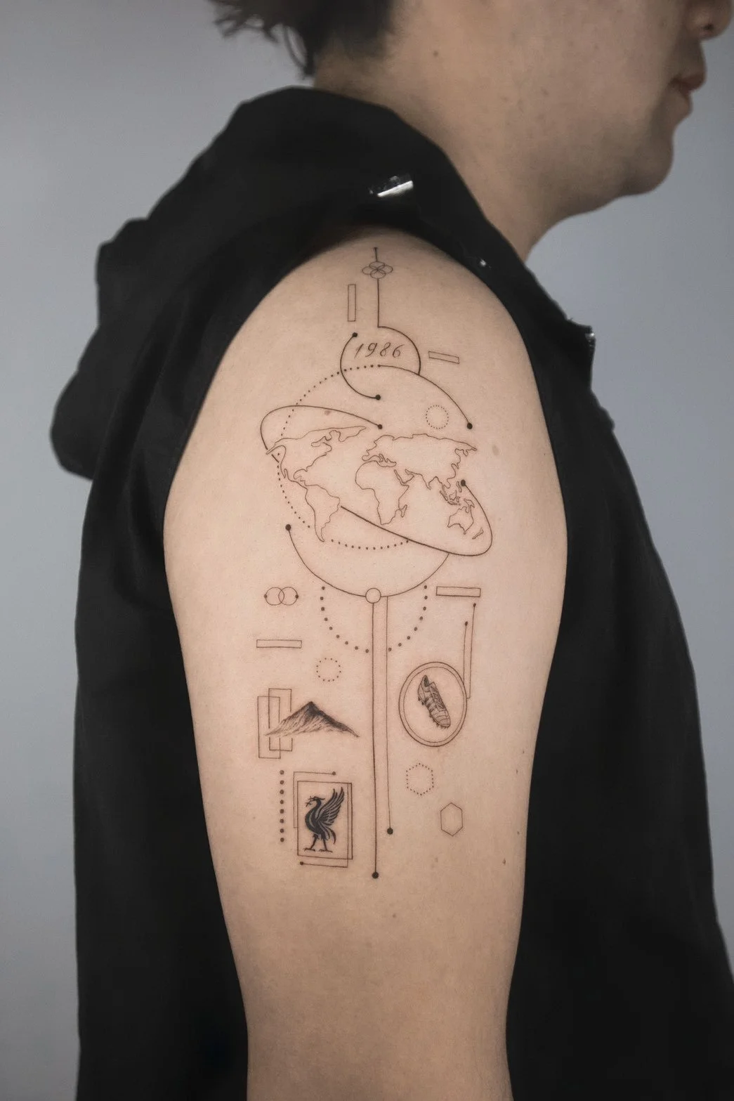 A tattoo on a person's upper arm featuring a minimalist line art design including a world map centered on Europe and Asia, geometric shapes, a mountain, a bird, and various small symbols, with the year 1986 inscribed.